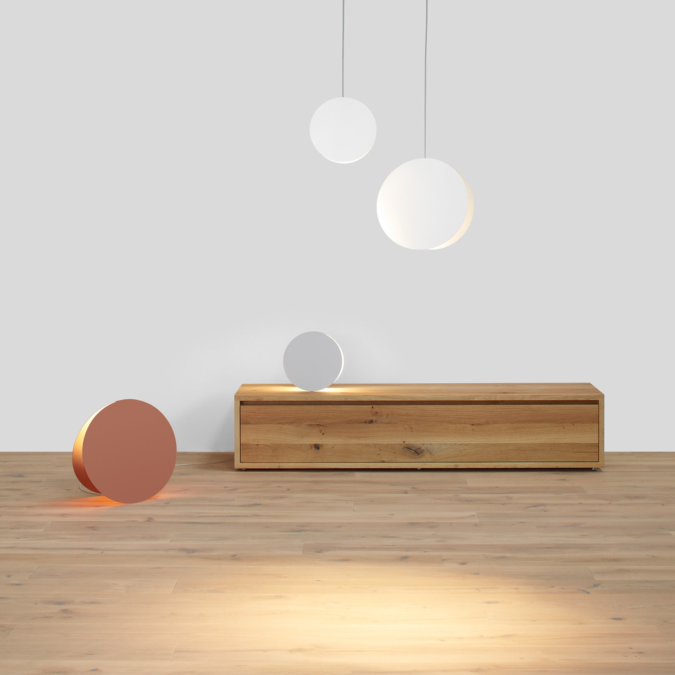A minimalist room showcases an E15 SB05 IMARI wooden bench with adjustable feet against a white wall. Three circular lamps hang above, casting soft light. A round, reddish-brown object on the light wood floor enhances the serene ambiance of solid wood elements.
