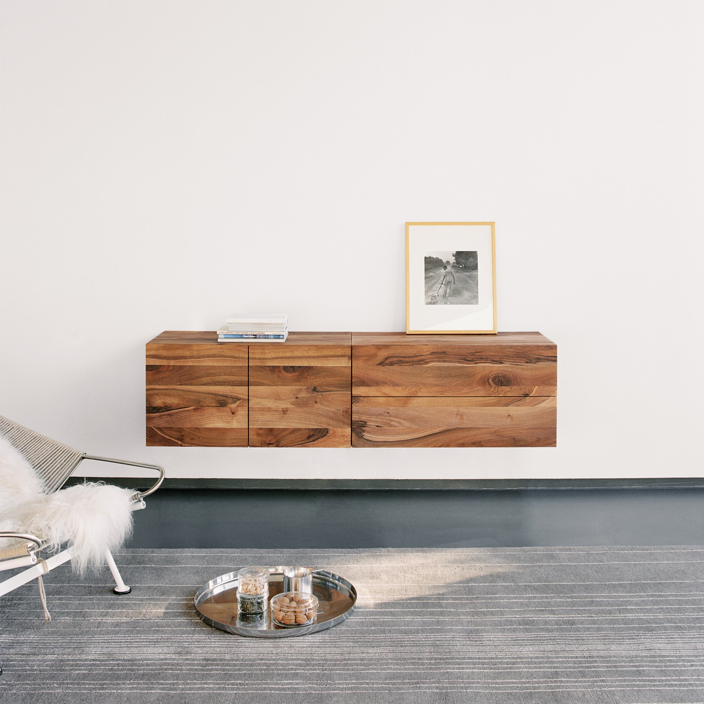 A minimalist room showcases E15s SB07 SHAHNAZ, a wall-mounted wooden cabinet on a white wall. It holds a framed photo and stacked books. A metal chair with a white cushion enhances the elegance, while a rug with cookies and dog treats completes the cozy scene.
