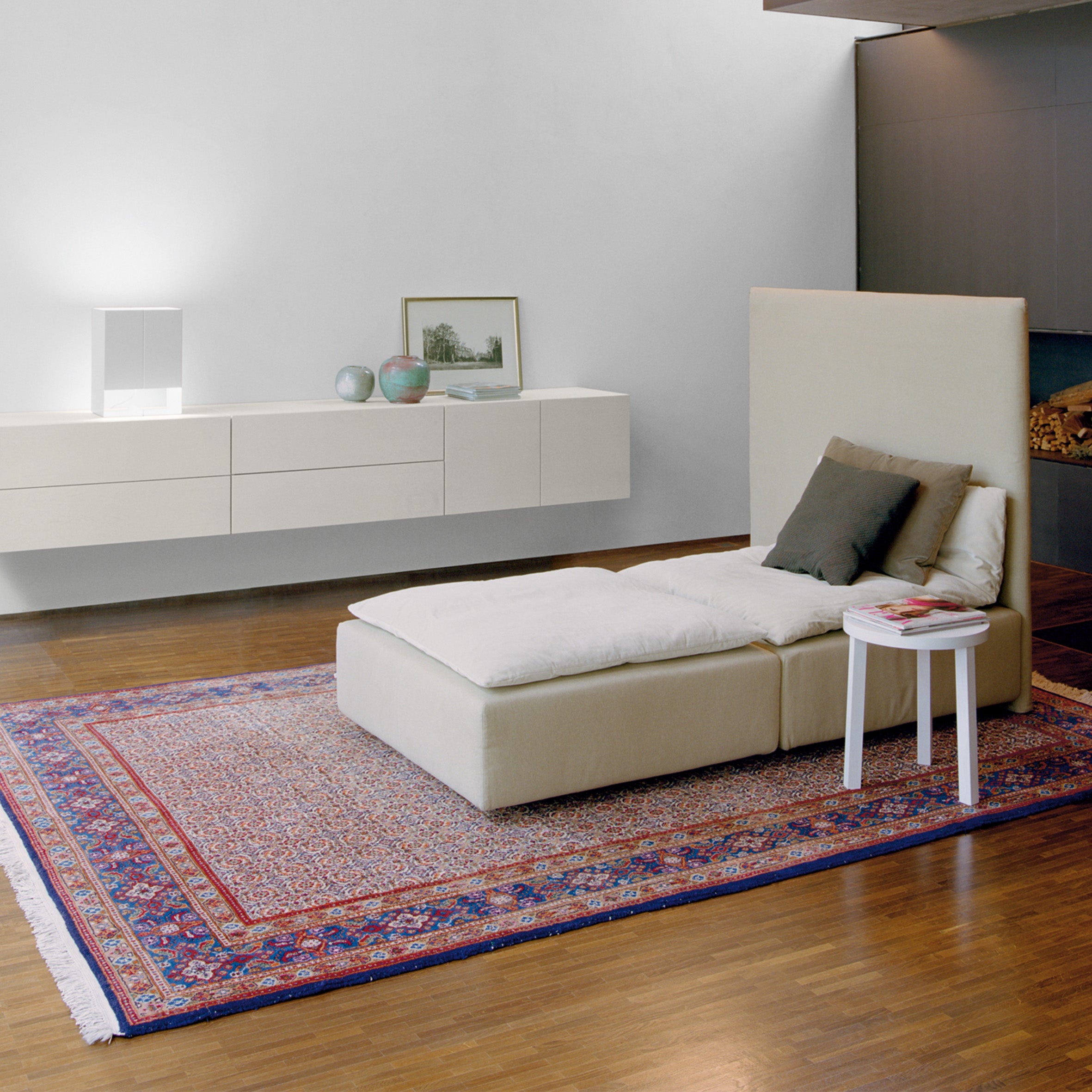 A minimalist living room features the beige E15 SB07 SHAHNAZ chaise lounge on a patterned rug, paired with an elegant wall-mounted console decorated with decor items. A white side table with a book adds charm, while wooden flooring and soft ambient lighting complete the sophisticated look.