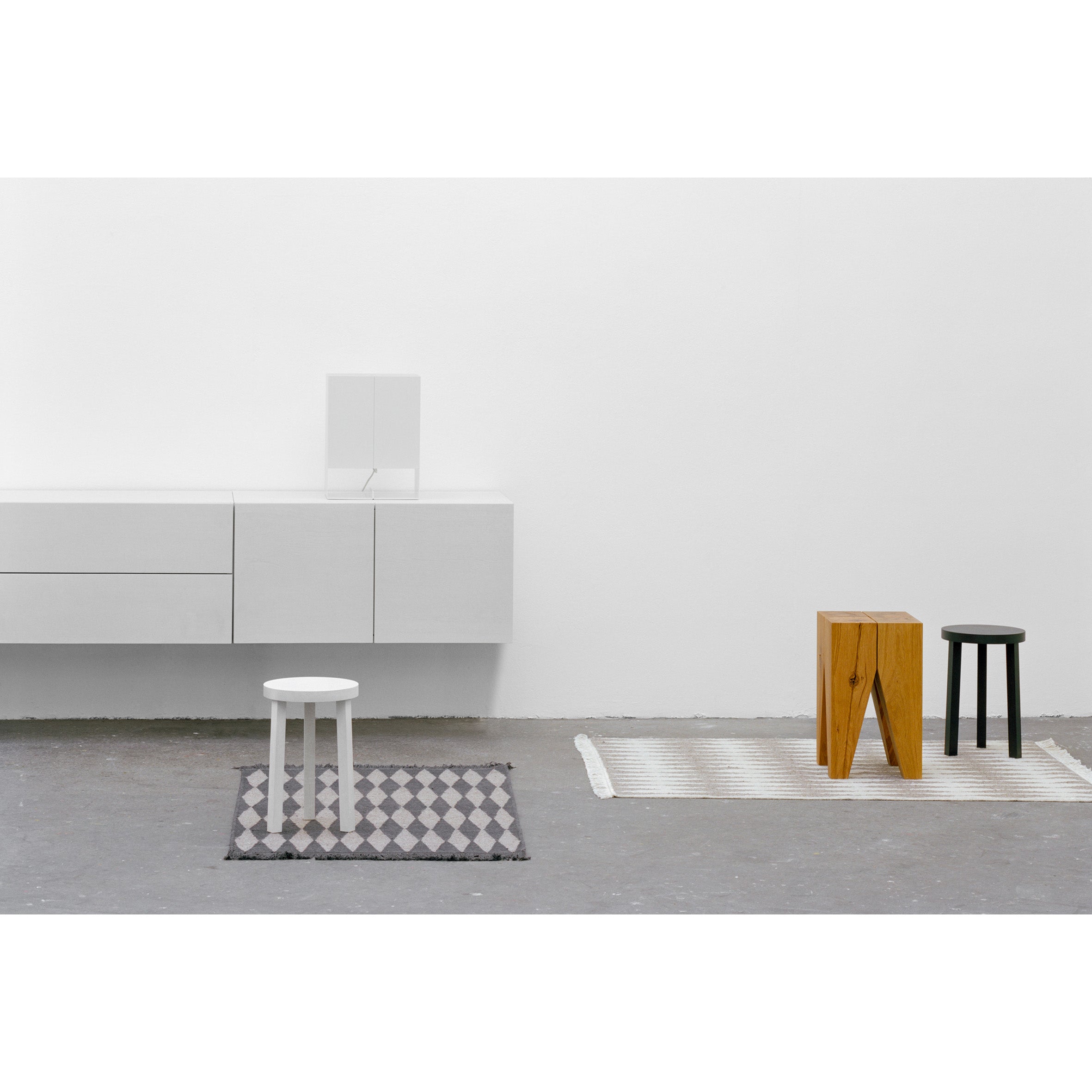 A minimalist room is elegantly designed with three stools: a white one on a gray patterned rug, a wooden one on a white rug, and a black one on a gray geometric-patterned rug. A wall-mounted E15 SB07 SHAHNAZ modular sideboard cabinet enhances the decor.