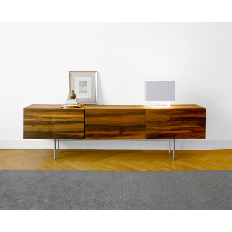 The SB07 SHAHNAZ by E15, with its modern and elegant design, stands against a white wall. It includes three cabinets and is topped with a framed picture and lamp. The floor features parquet patterning, accented by a gray rug.