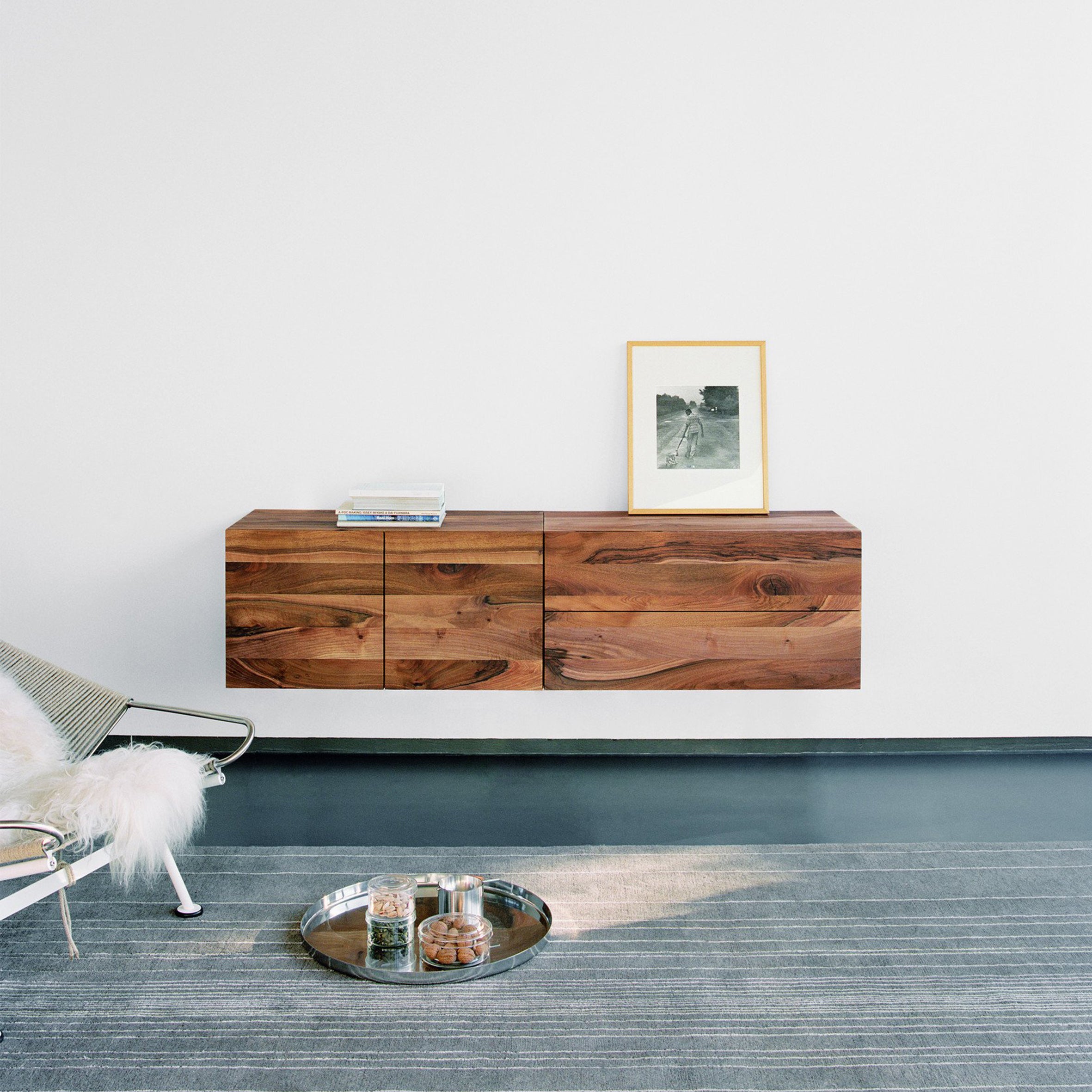 A minimalist living space showcases the E15 SB08 MAHNAZ, a floating wooden cabinet with chamfered edges against a white wall. It holds a framed photo and books. Nearby, a chair with a fur throw sits beside a tray with candles and snacks on a striped rug.
