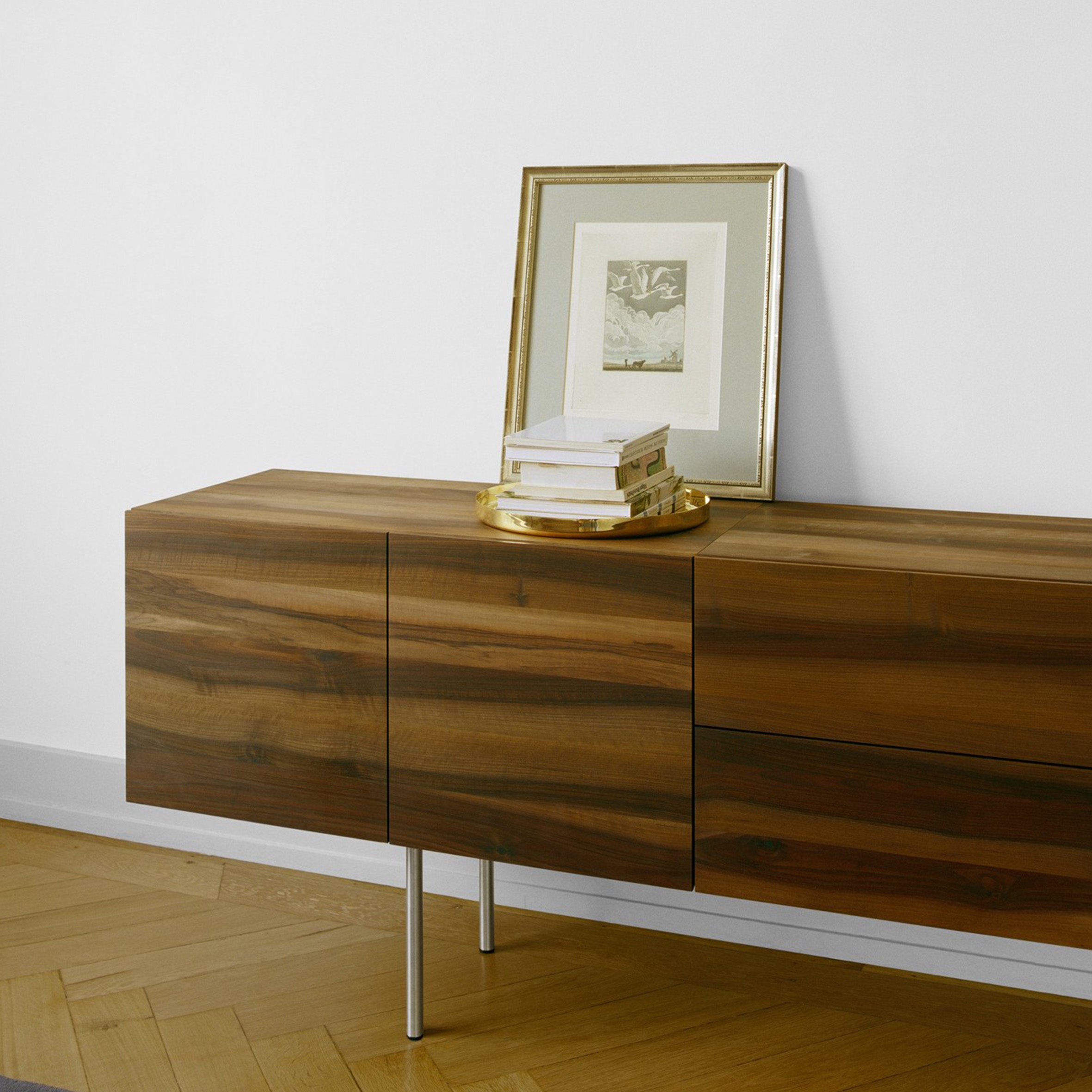 The SB08 MAHNAZ sideboard by E15, featuring a smooth natural finish and elegant handles, stands against a white wall. A framed picture, books, and a golden tray are neatly arranged on top. The floor showcases a wooden diagonal pattern.
