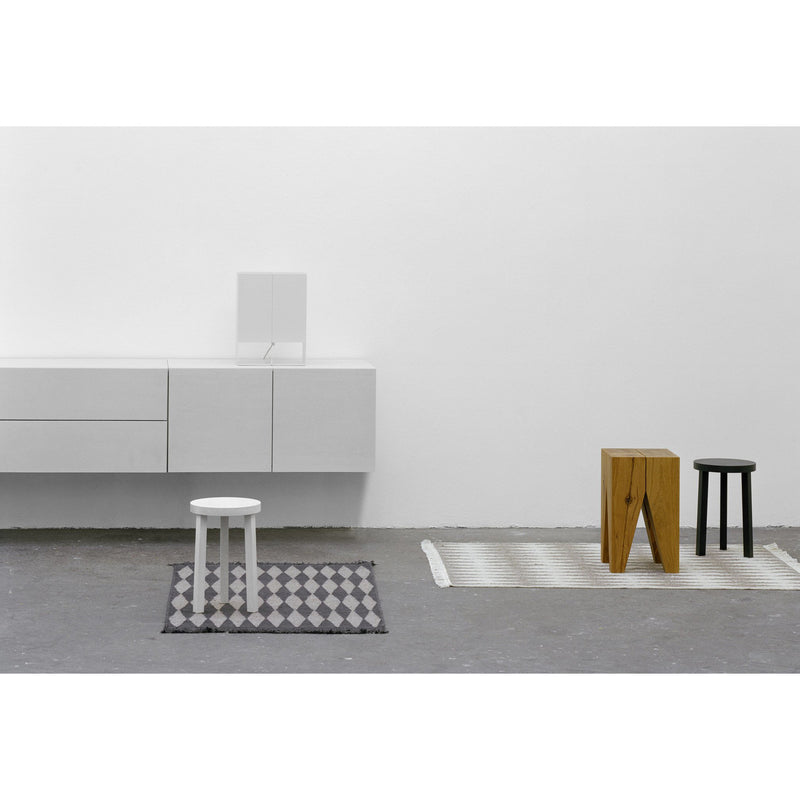 A minimalist room showcases the E15 SB08 MAHNAZ white wall-mounted cabinet with an elegant handle, two small patterned rugs, and three stools—white, wooden, and black. The floor is concrete with a small mirror leaning above the cabinet.
