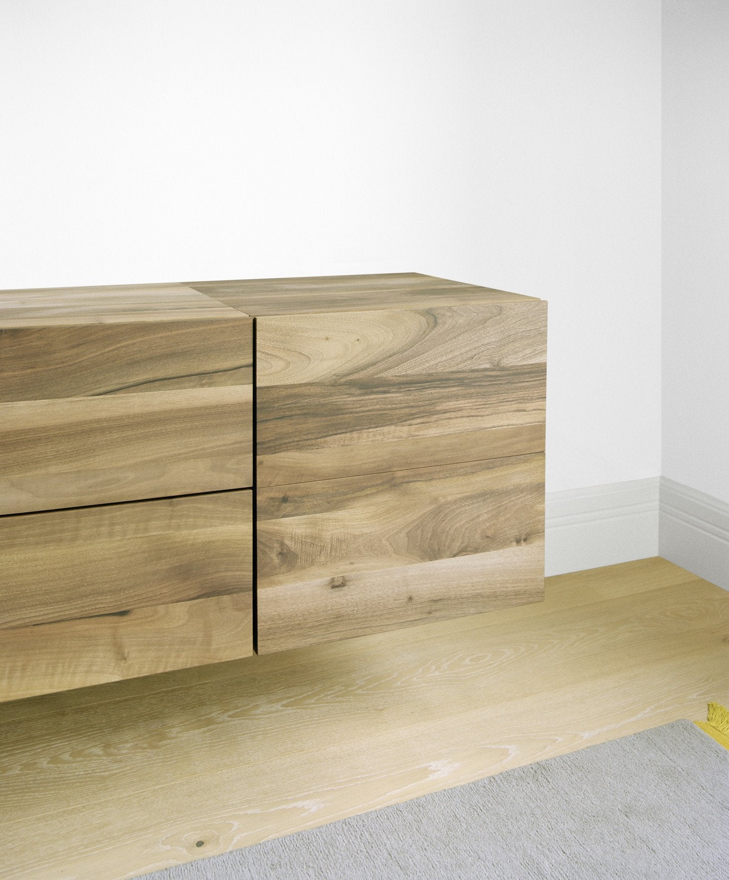 The SB10 ARAQ by E15 is a floating wooden cabinet with a natural grain finish, set against a white wall. This solid wood sideboard, featuring two clean-lined segments, is mounted above a light wood floor partially covered by a gray rug.