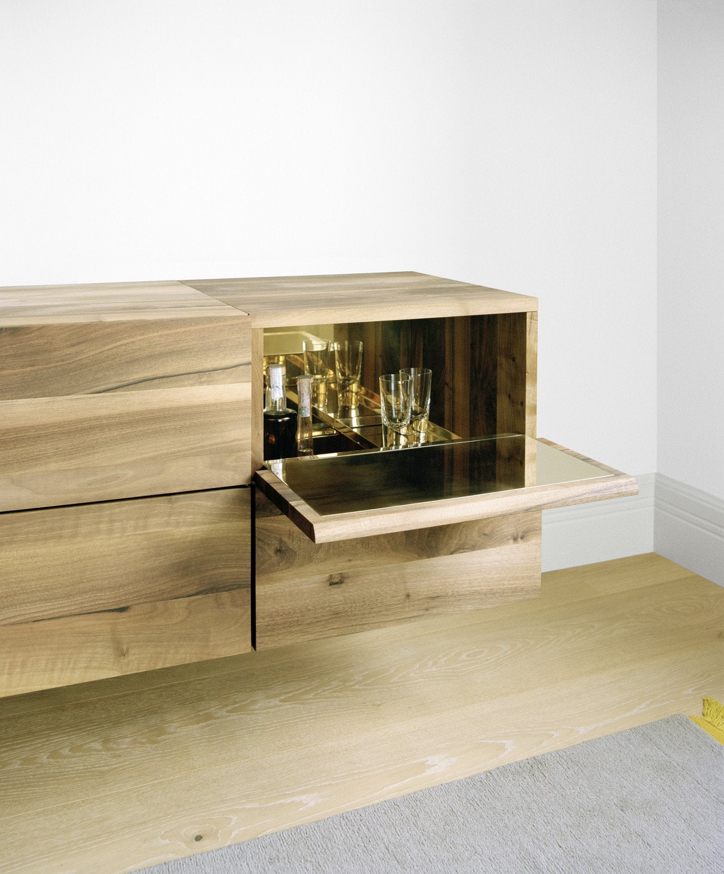 The SB10 ARAQ by E15 is a wooden sideboard with a sleek design that doubles as a bar cabinet. It features an open compartment for glassware and bottles, with a mirrored shelf inside to reflect light. The light wood flooring enhances its elegance, complementing the polished brass interior accents.