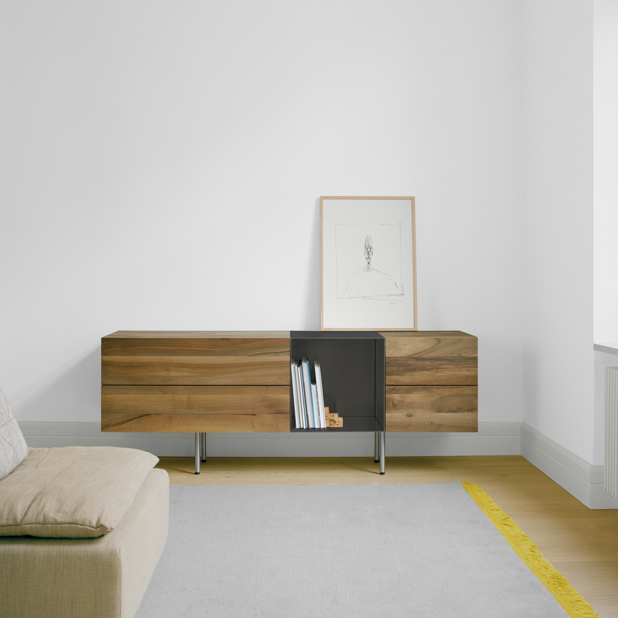 A contemporary living room highlights E15s SB11 DARA wooden sideboard, with an open shelf displaying books. A framed sketch adorns the wall above. A beige sofa complements the light gray rug on the wooden floor, crafting an inviting ambiance.
