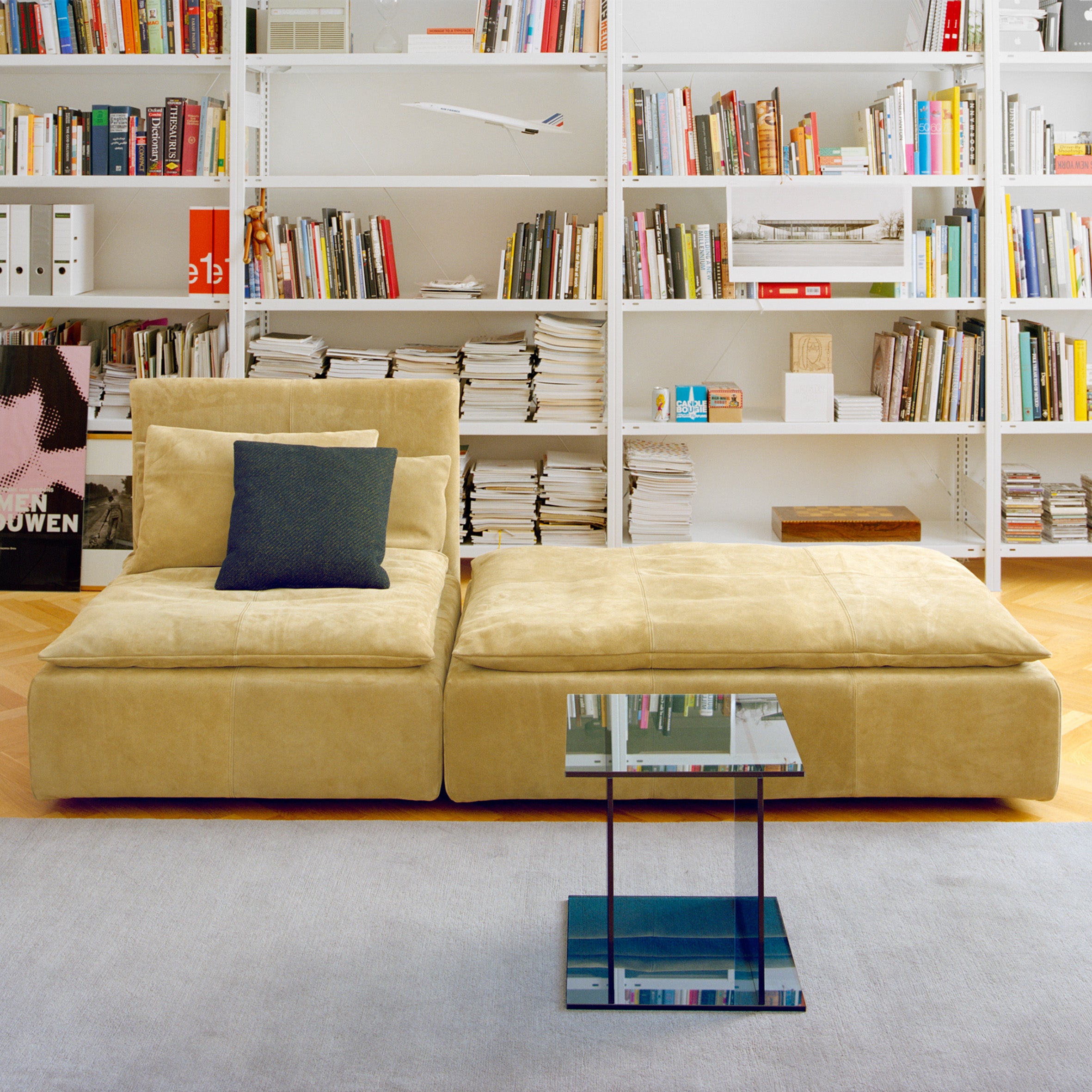 A modern living room showcases an E15 SF03 SHIRAZ sofa with a beige sectional and dark square pillow. A small glass coffee table is in front, while white shelves in the back display books, magazines, and decorative items.