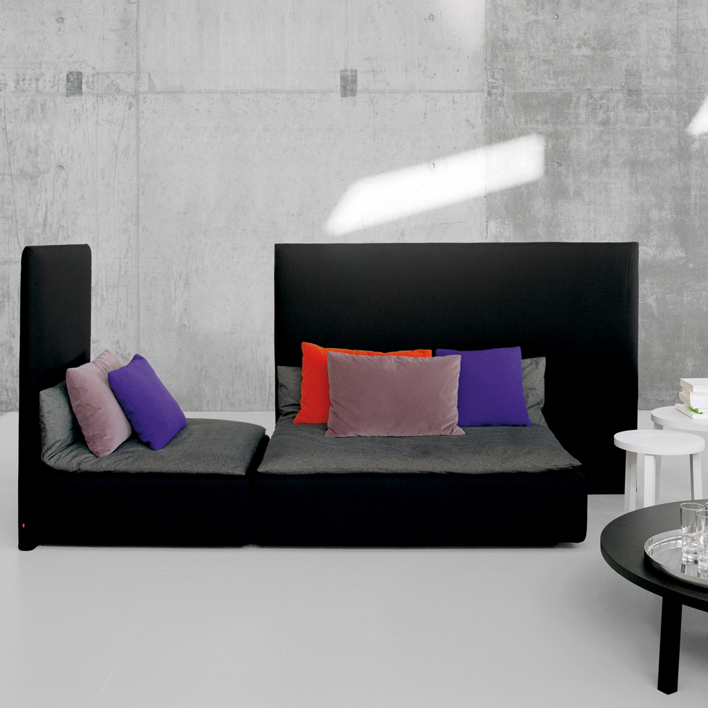 The contemporary living room features the SF03 SHIRAZ sofa by E15, a black modular design with a high backrest, embellished with red, purple, and gray cushions. It is flanked by two white side tables loaded with books against concrete walls and gray floors.