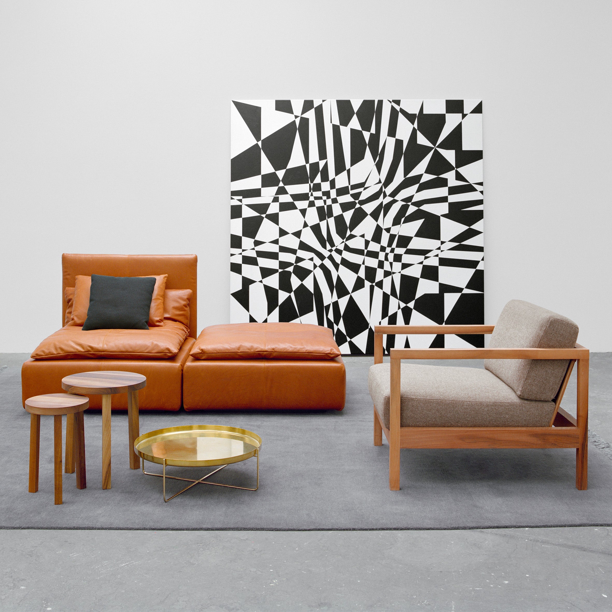 A modern living room includes a brown leather chair and ottoman, a wooden armchair with gray cushions, two circular wooden tables, and the E15 SF03 SHIRAZ sofa. This setup is beneath a large black-and-white abstract art piece on a gray carpet.