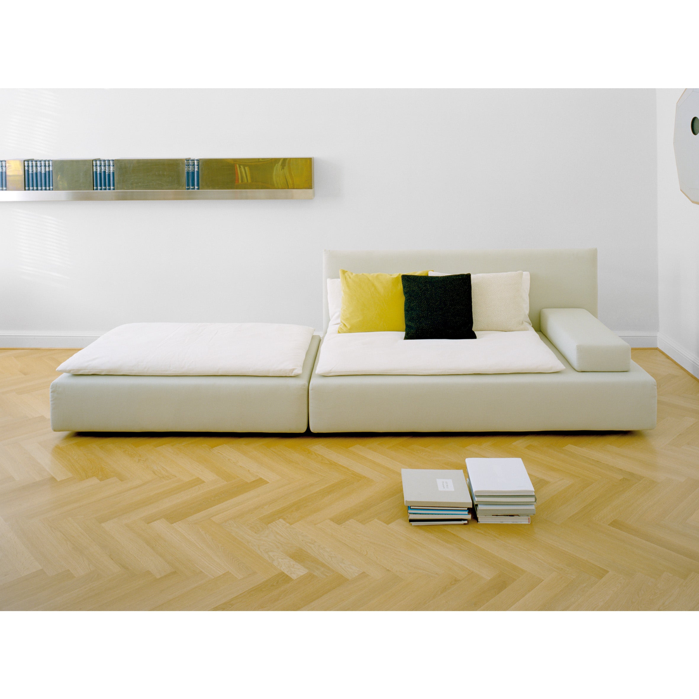 A minimalist living room features the SF03 SHIRAZ sofa by E15, a modern, white modular piece with yellow and black cushions. Above it, a sleek wall shelf holds books, while more are stacked on the light wood herringbone floor, enhancing this flexible system of contemporary comfort.