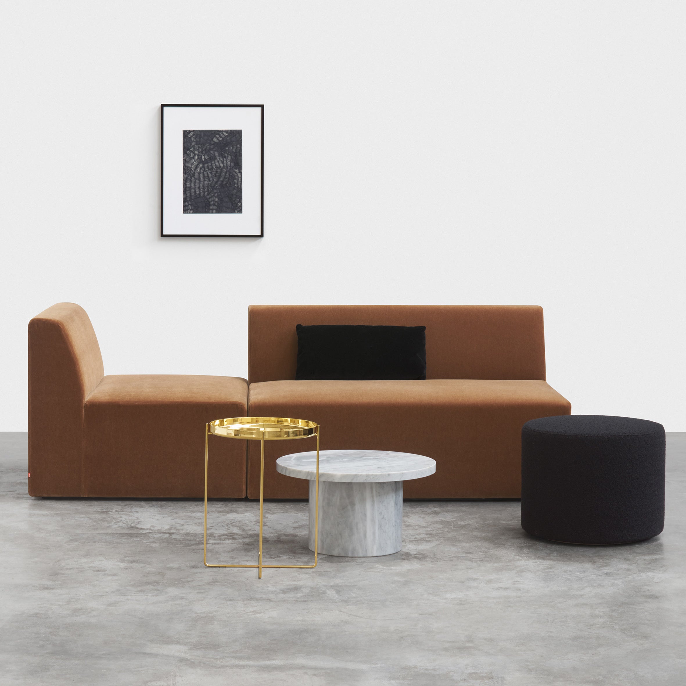 A minimalist living room showcases a brown SF06 KERMAN POUF by E15 with black cushions. Two coffee tables—one gold with a glass top and one round marble—enhance the space. Nearby, a black ottoman offers extra seating, while framed abstract artwork adorns the white wall.