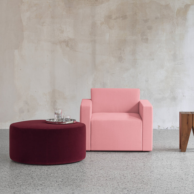 A modern minimalist interior includes a pink armchair next to the SF06 KERMAN POUF by E15, topped with a silver tray and glassware. The setting boasts a textured gray wall, speckled floor, and versatile seating modules on the right with a wooden table featuring removable covers.