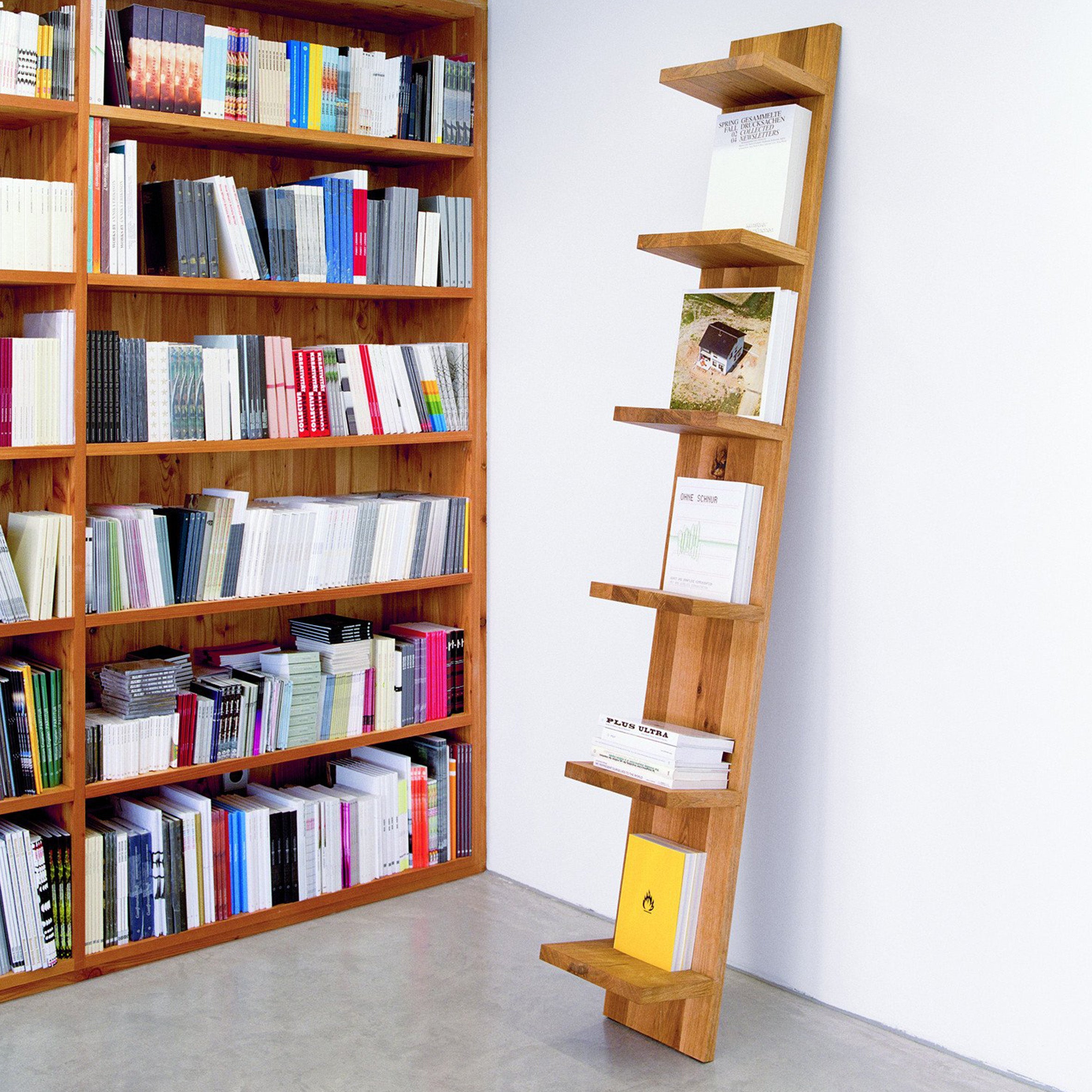 A minimalist leaning bookshelf holds a few books against a white wall. To the left, an E15 SH01 MATE solid wood shelf is filled with colorful books, creating a vivid contrast. The floor is light gray concrete.