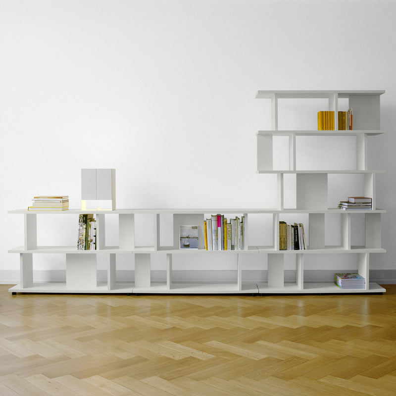 The SH05 ARIE by E15 is a minimalist white bookshelf with an asymmetrical design, featuring vertical and horizontal sections. It stylishly holds books, a lamp, and decor items against a light wood floor and white backdrop, reflecting E15 Designer inspirations.