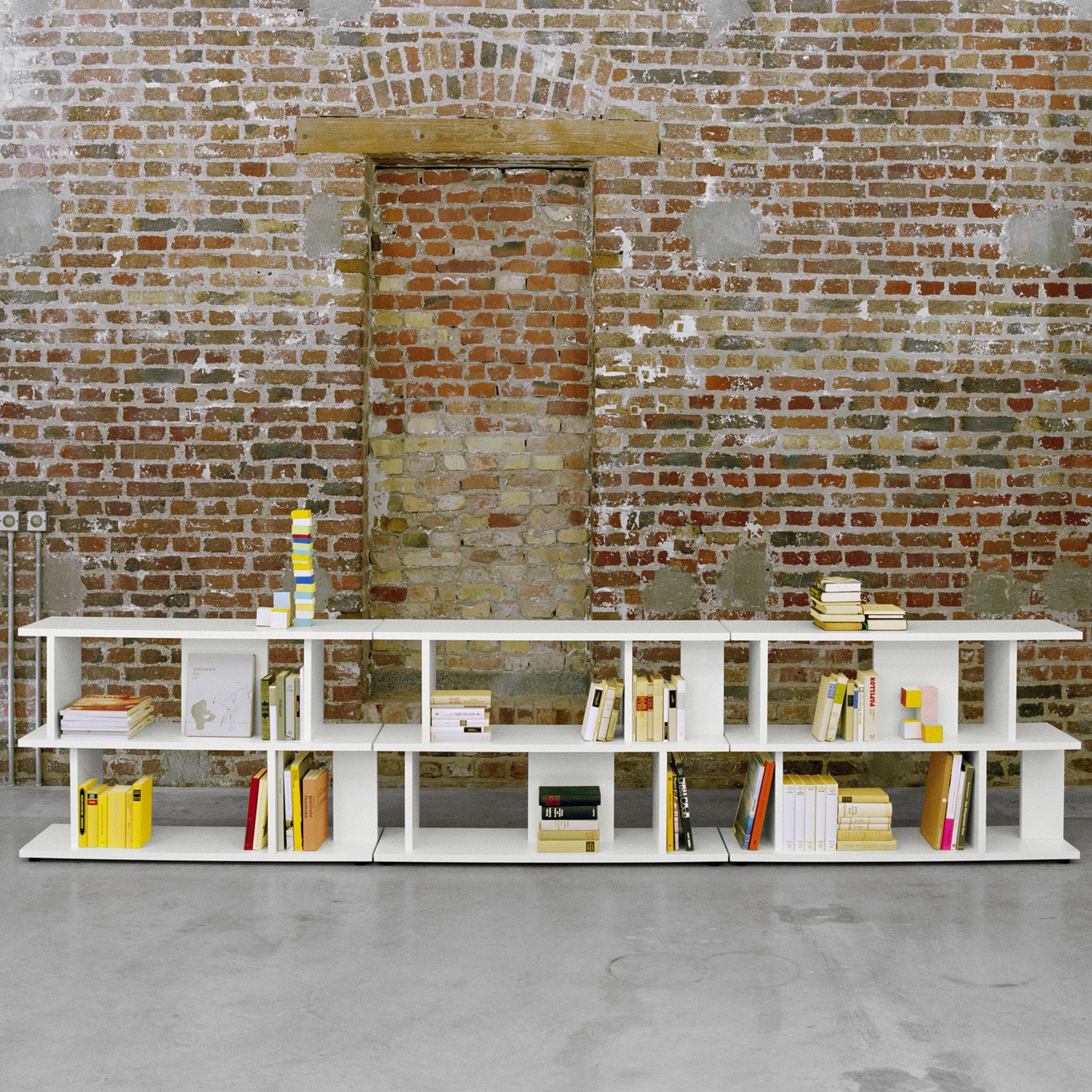 The SH05 ARIE by E15, a modern white bookshelf, is set against an exposed brick wall, filled with vertically and horizontally arranged books. An archway behind serves as a shelving wall, while the floor exhibits smooth concrete finish.
