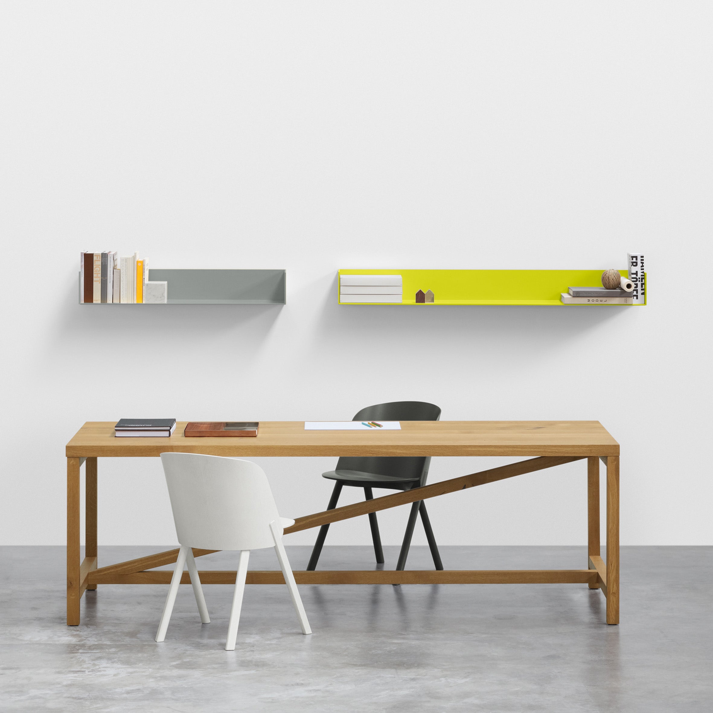 Create a minimalist office setup featuring the E15 SH06 PROFIL wooden desk, two modern chairs in white and dark green. Use a wall-mounted shelf with a yellow back to display books and decor, all against a white wall with minimal art.