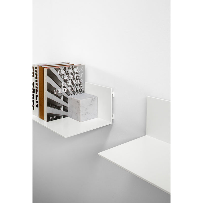 A minimalist white wall features two sleek, wall-mounted shelves from E15. The SH06 PROFIL holds stacked books, including Architect Designs Modern Living, while the adjacent aluminum shelf remains empty and clean, highlighting simplicity and modern design as minimal art.