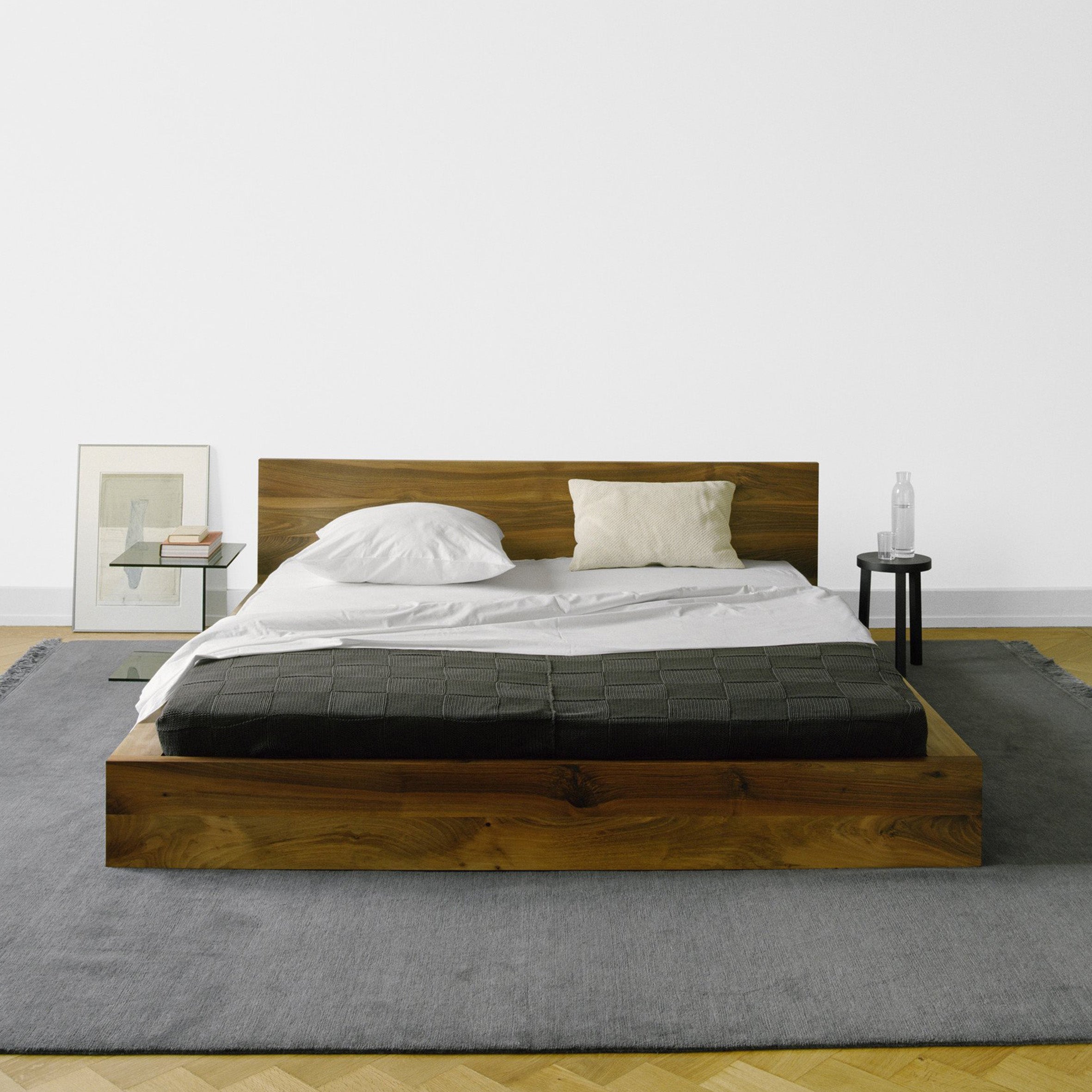 A minimalist bedroom features an E15 SL02 MO solid wood bed with white sheets, a black blanket, and a single pillow. A small black side table holds a water bottle and glass. A framed picture and book rest on a gray rug beside the bed.