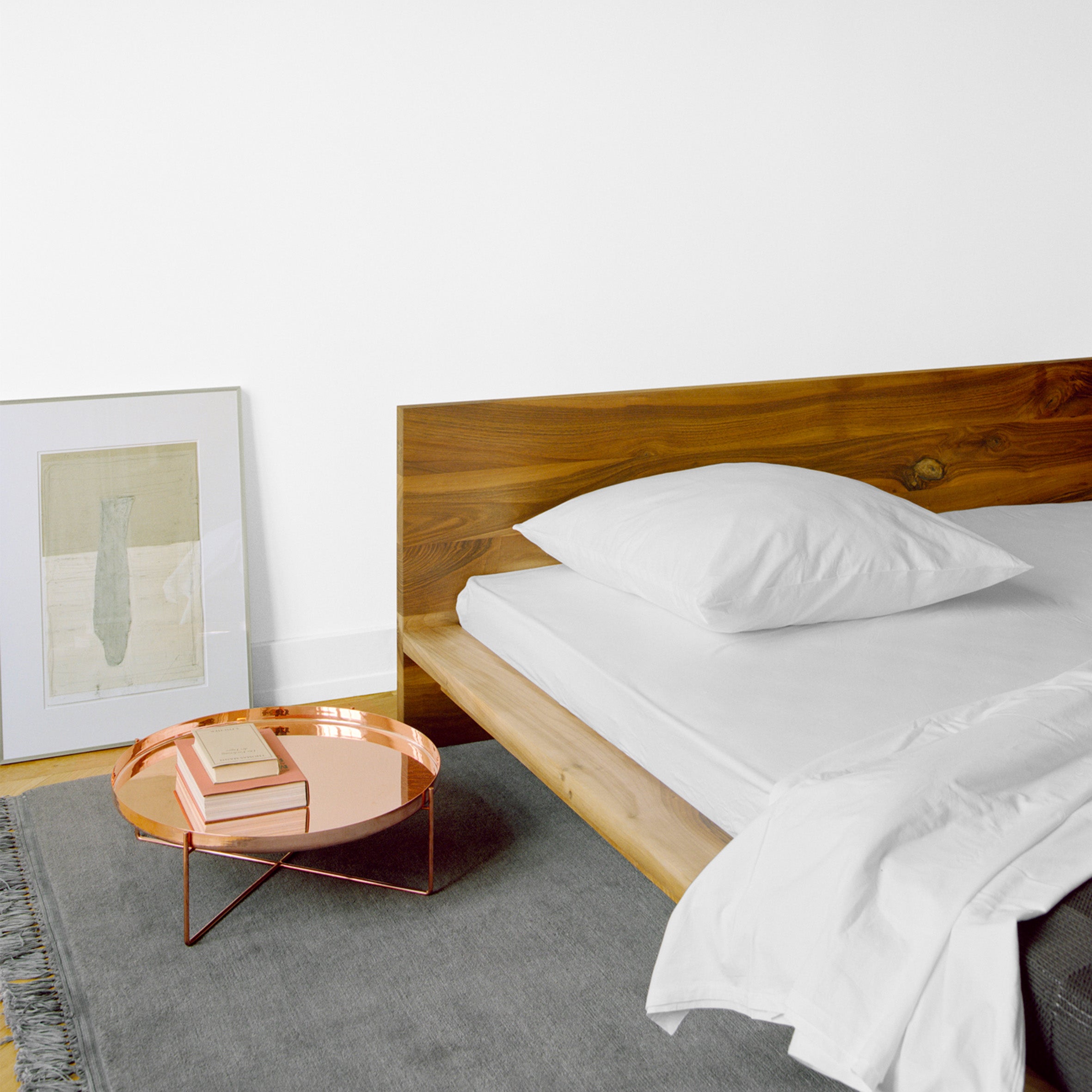 A minimalist bedroom showcases the E15 SL02 MO solid wood bed adorned with white sheets and a single pillow. Beside it, a copper tray table holds books. A framed abstract artwork rests against the white wall, while a gray rug drapes the wooden floor.