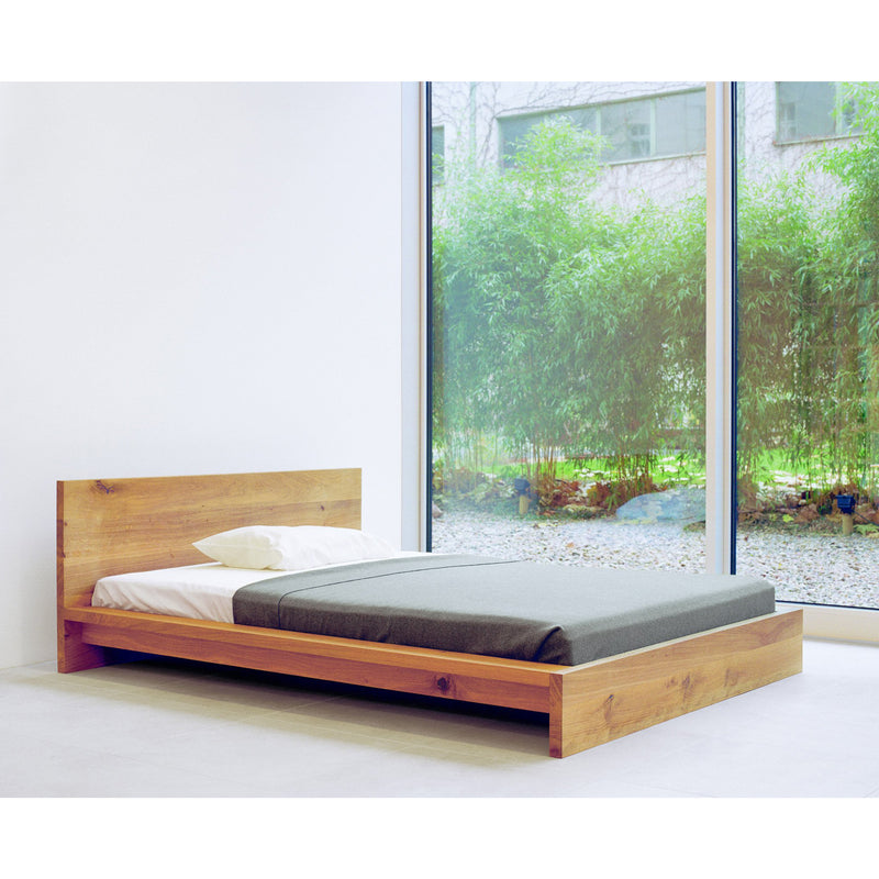A minimalist bedroom showcases the SL02 MO by E15, a low, solid wood platform bed adorned with a gray blanket and white pillow. Large windows reveal bamboo plants in the garden, infusing natural light and serenity into the space.