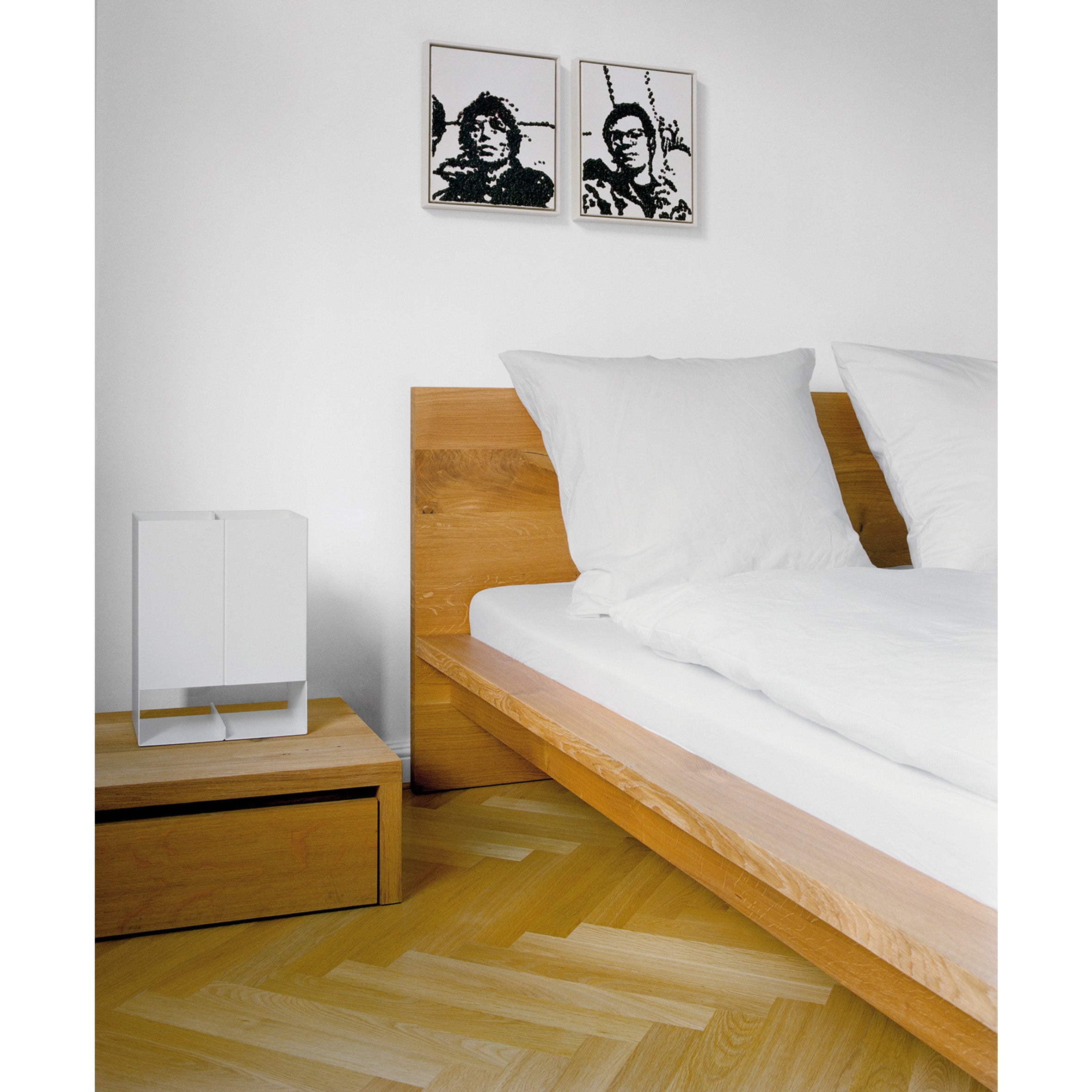 The minimalist bedroom showcases a designer E15 solid wood bed with white bedding. A small white cabinet sits on the wooden nightstand next to the SL02 MO bed, while two monochrome portraits adorn the white wall above, adding to the rooms elegant simplicity.