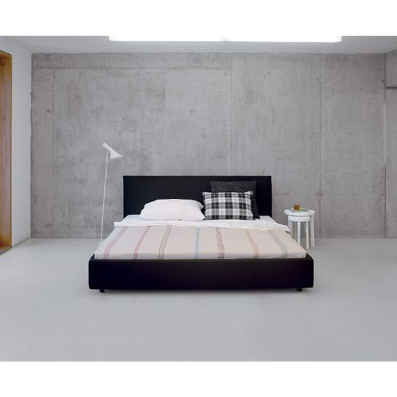 A minimalist bedroom showcases the E15 SL05 PARDIS upholstered bed with a black frame, neatly made with striped bedding, two pillows, and a checkered cushion. A white floor lamp is on the left, a small round table on the right, and concrete walls enhancing the sleek design.