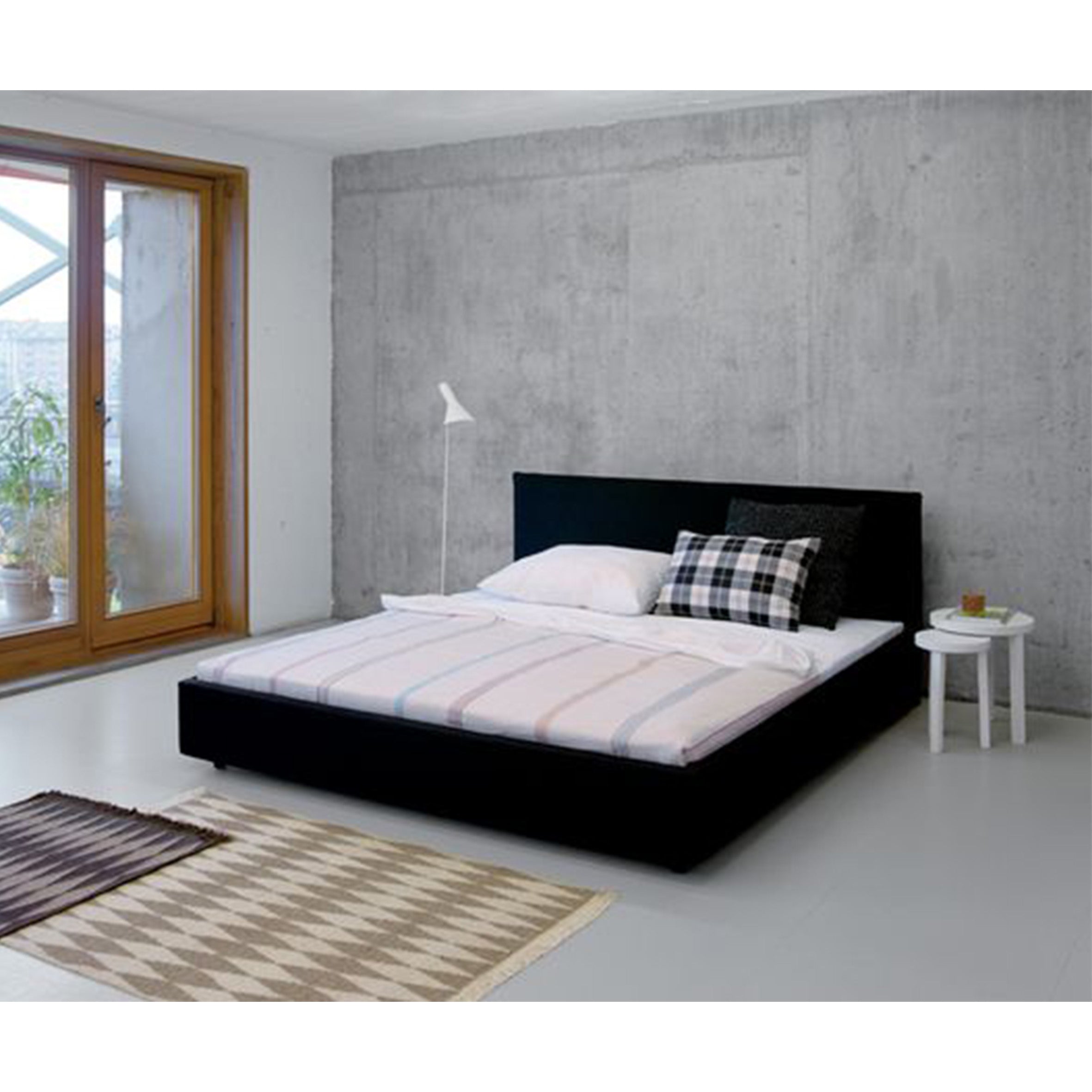 A minimalist bedroom with a low black E15 SL05 PARDIS bedframe against a concrete-style wall, adorned with a plaid pillow and striped quilt. Nearby are a white floor lamp and side table holding a book. To the left is a large wooden-framed window overlooking lush plants.
