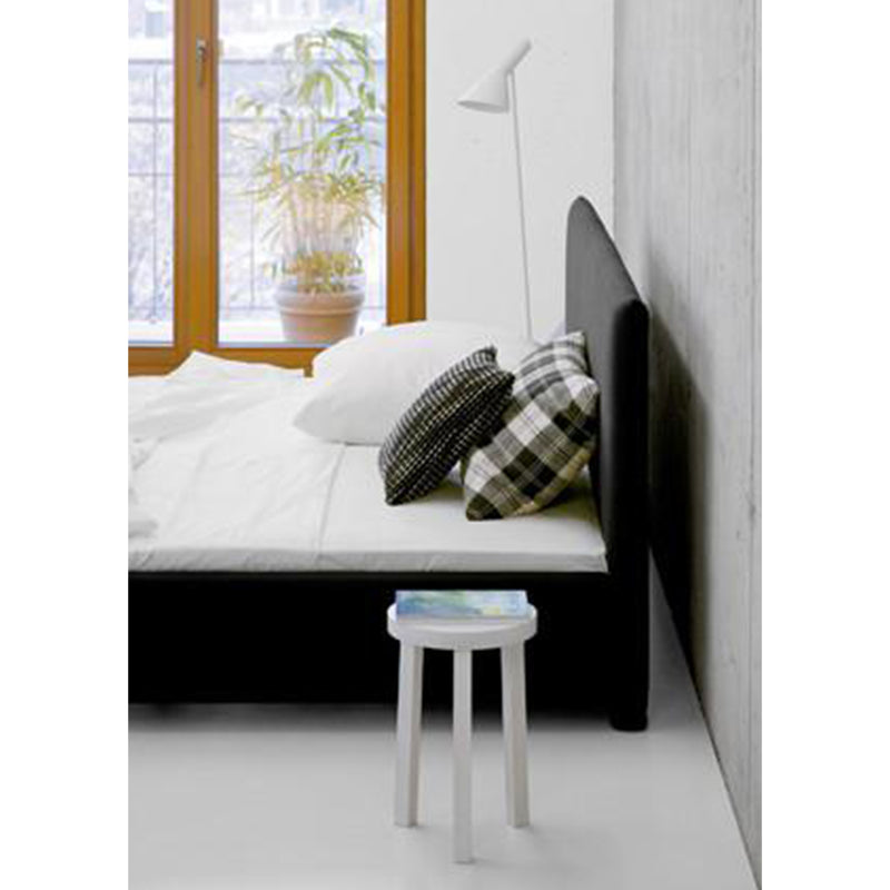 A minimalist bedroom showcases a sleek E15 SL05 PARDIS bed adorned with patterned pillows and white bedding. A small stool with a book sits at the foot, while a white floor lamp stands nearby. A bright window and potted plant complete this serene space.