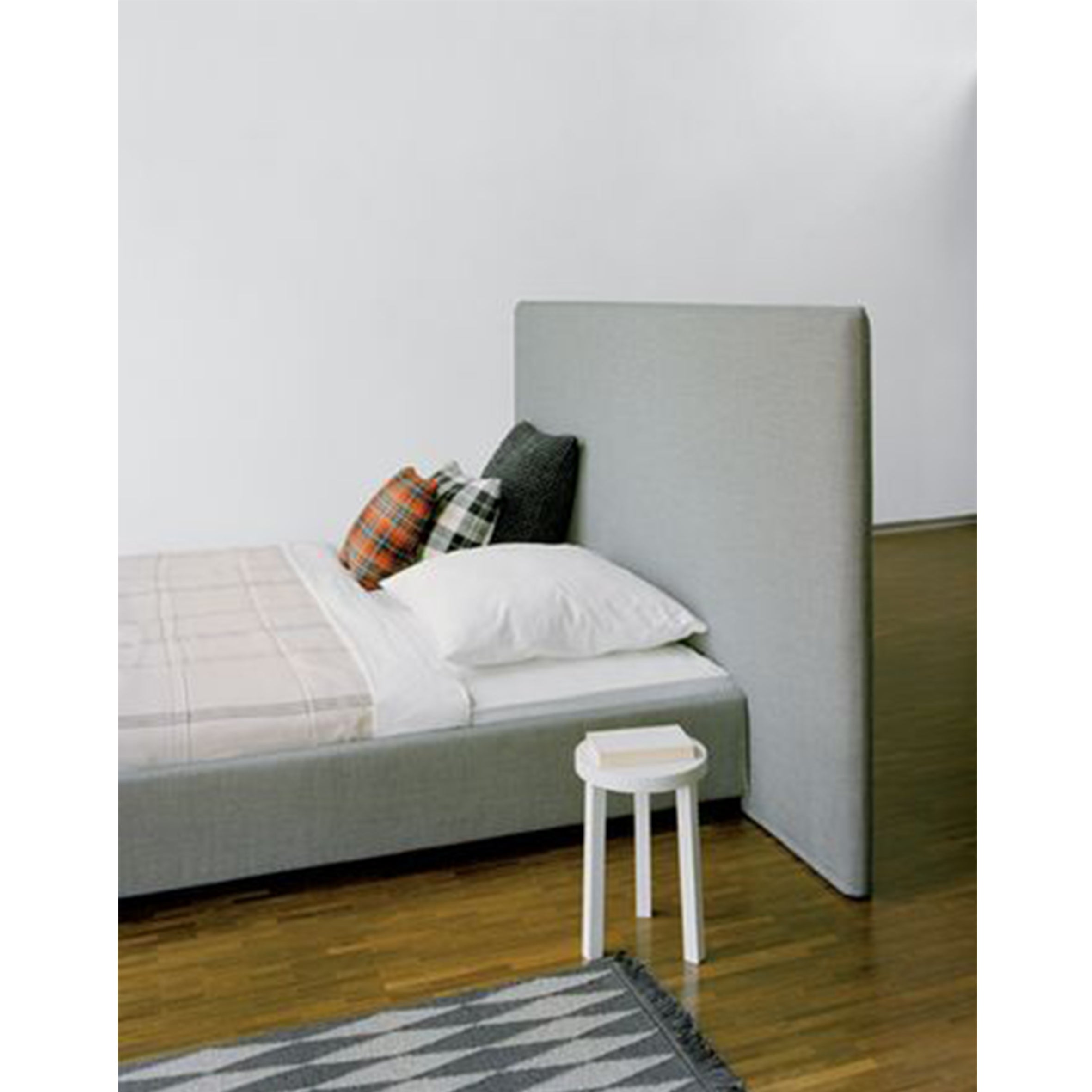 This minimalist bedroom features E15s SL05 PARDIS upholstered bed with white and plaid pillows and a light brown blanket. A small white stool complements the wooden floor, geometric-patterned rug, modern headboard, and plain white walls.