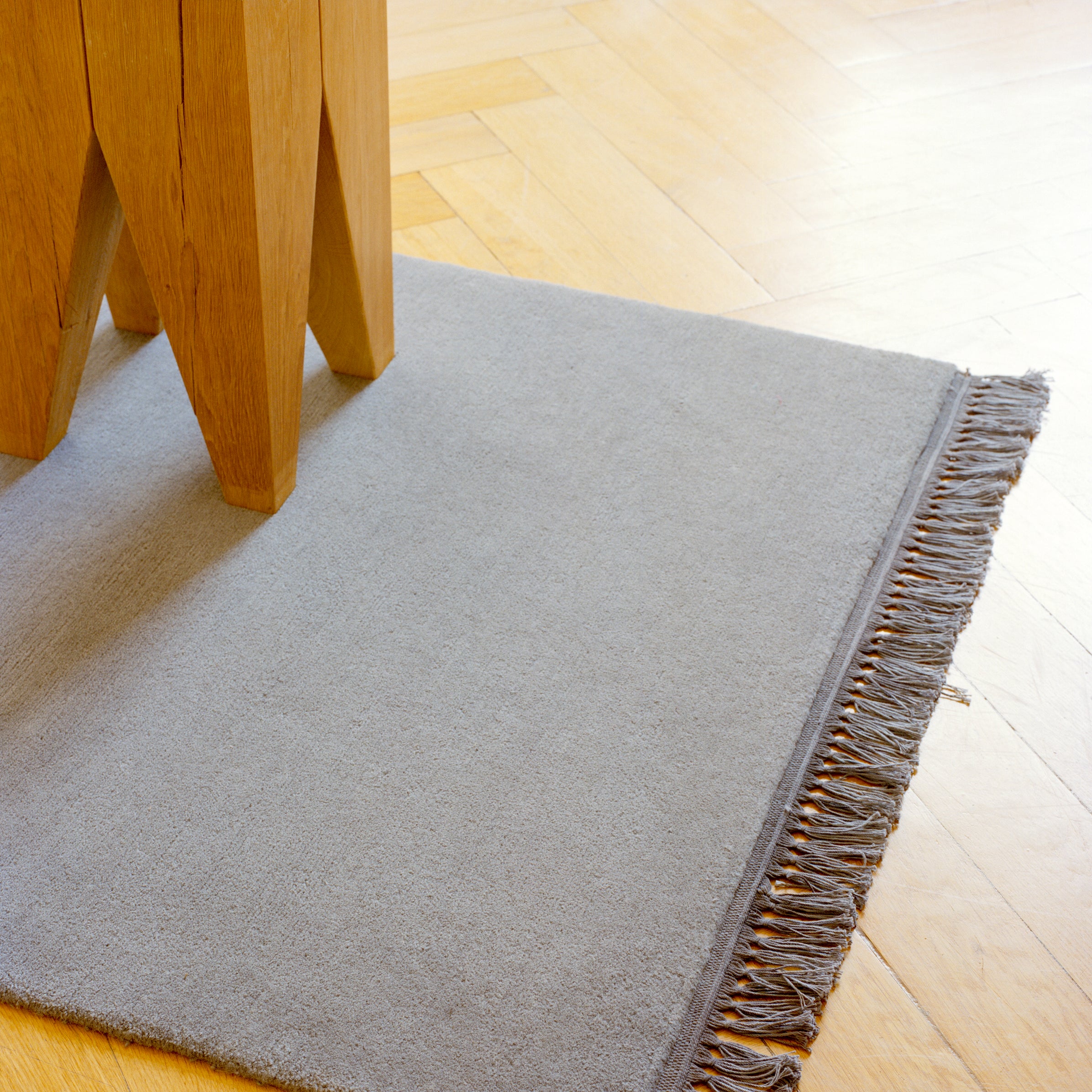 A light gray fringed rug corner lies under an E15 wooden table on a light wood floor in a herringbone pattern. An elegant ST04 BACKENZAHN solid wood stool stands beside it, all illuminated by natural light.
