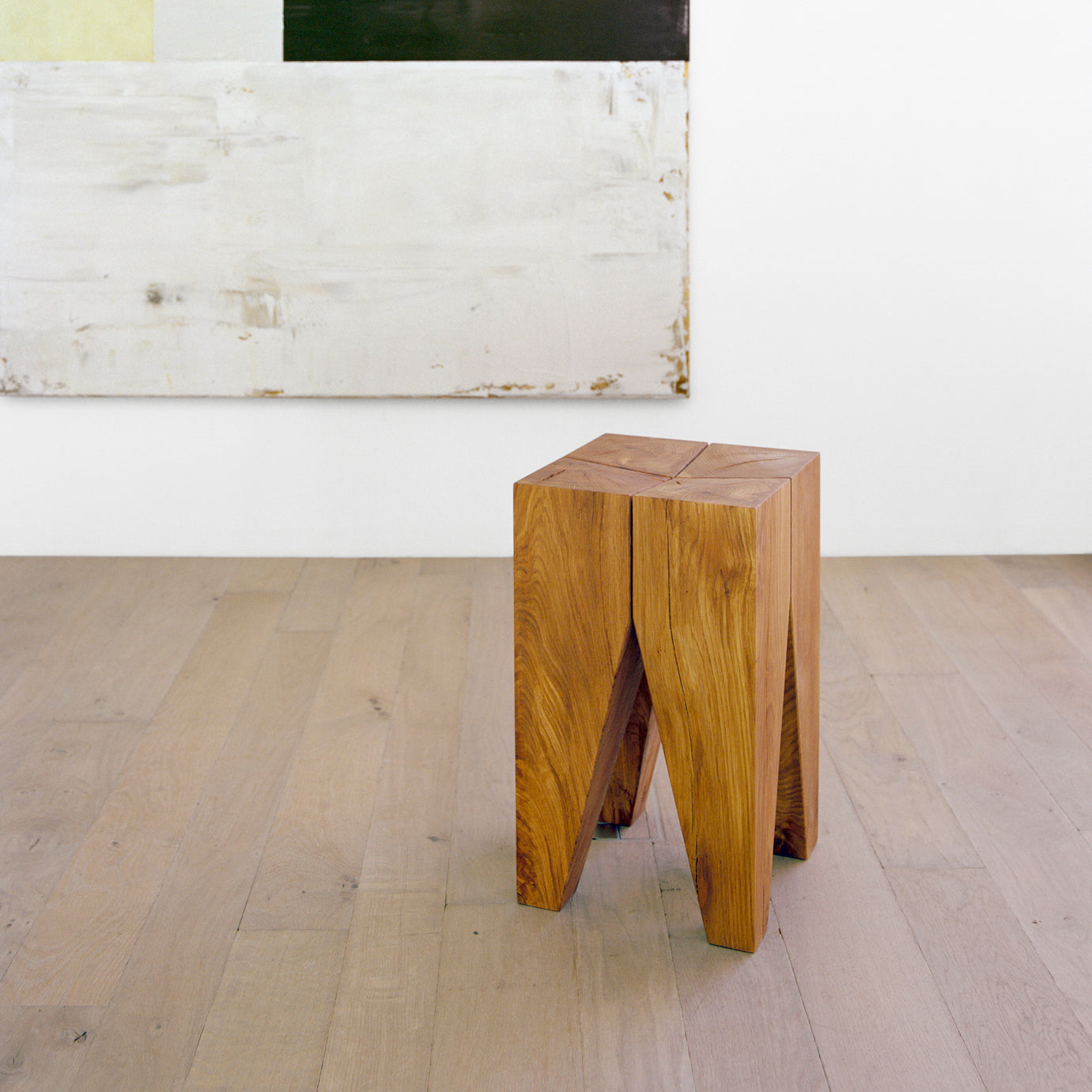 An E15 ST04 BACKENZAHN stool with a geometric design sits on a light wood floor. A large abstract painting, showcasing black, white, and light gray rectangles, hangs on the white wall behind it.