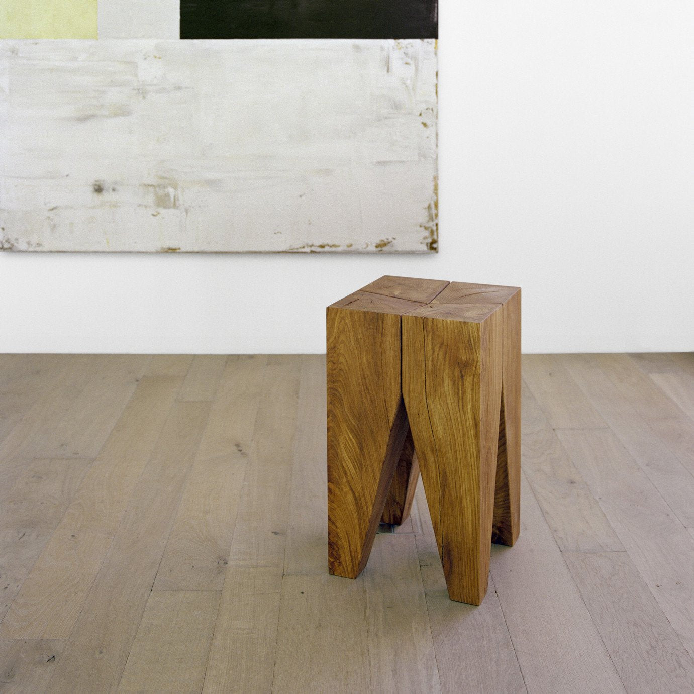 An E15 ST04 BACKENZAHN side table with its distinctive geometric design rests on a light wooden floor. In the background, a large abstract painting featuring white, black, and beige blocks adorns a simple white wall.