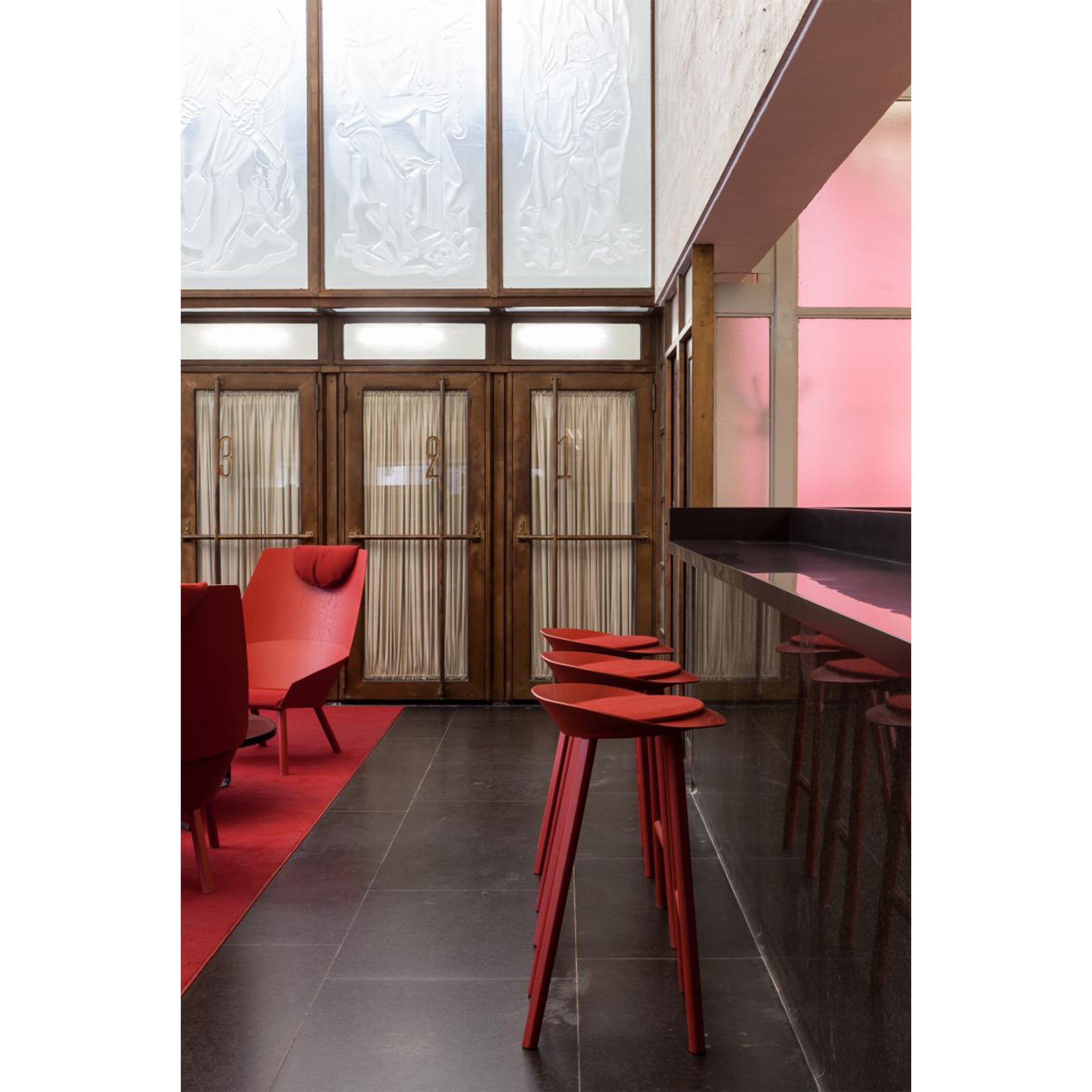 A modern interior featuring red chairs and ST10 JEAN bar stools from E15, complemented by a sleek black counter and dark tiled flooring. Tall oak veneered plywood doors with decorative panels are highlighted by warm pink lighting.