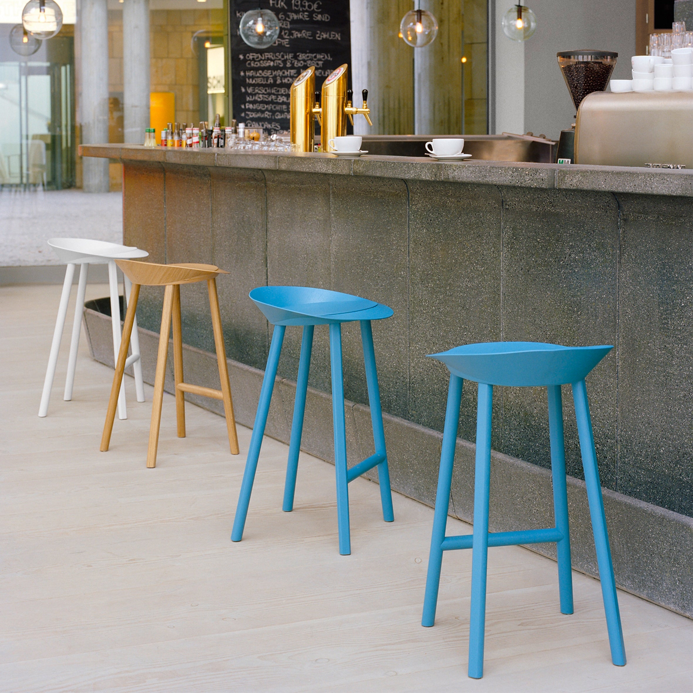 A modern bar showcases vibrant bar stools in blue, brown, and white. The E15 ST10 JEAN model stands out with its industrial design, perfectly complementing the warm oak veneered plywood counter. Coffee cups appear against a blurred background in the well-lit setting.