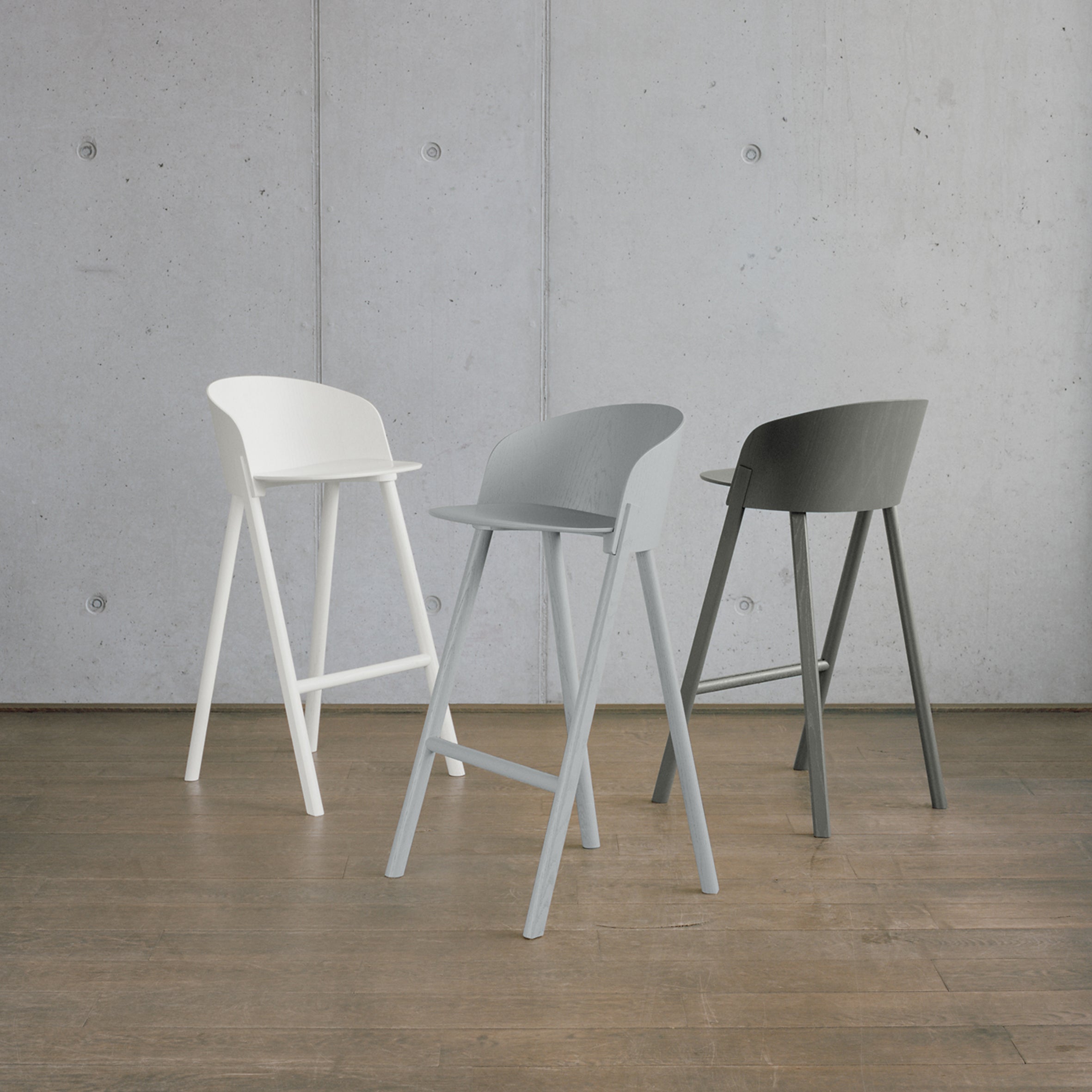 Three E15 ST12 OTHER modern bar stools in white, light gray, and dark gray stand on a wooden floor against a concrete wall. They feature sleek curved backrests and four-legged frames, blending style with comfort.