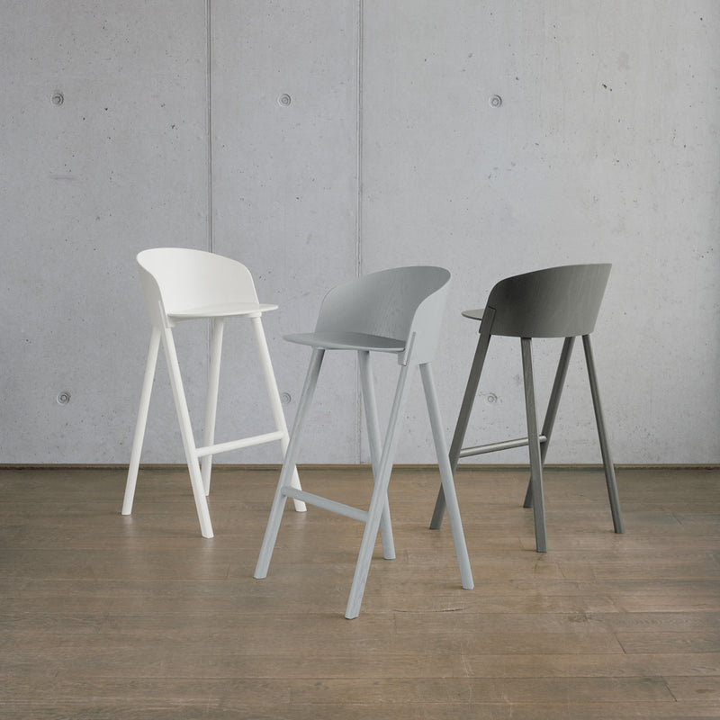 Three sleek and minimalist ST12 OTHER bar stools from E15s Stefan Diez seating series, featuring shaped backrests and footrests, are arranged on a wooden floor against a concrete wall in white, light gray, and dark gray.