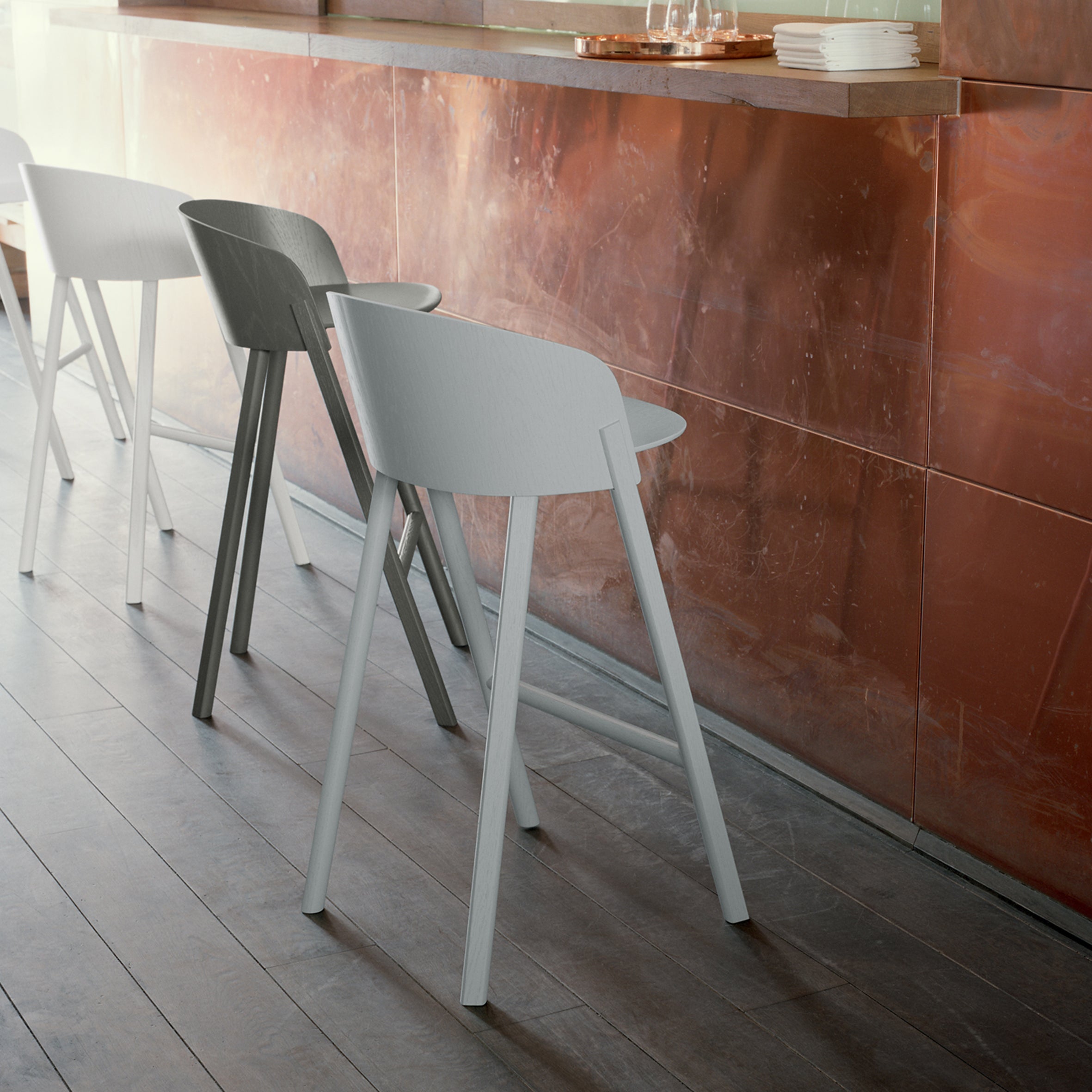 A row of E15s ST12 OTHER modern chairs with backrests lines up against a textured copper wall. Their sleek design and subtle upholstery contrast with the walls warm tones, while the wooden floor enhances this stylish, comfortable setting.
