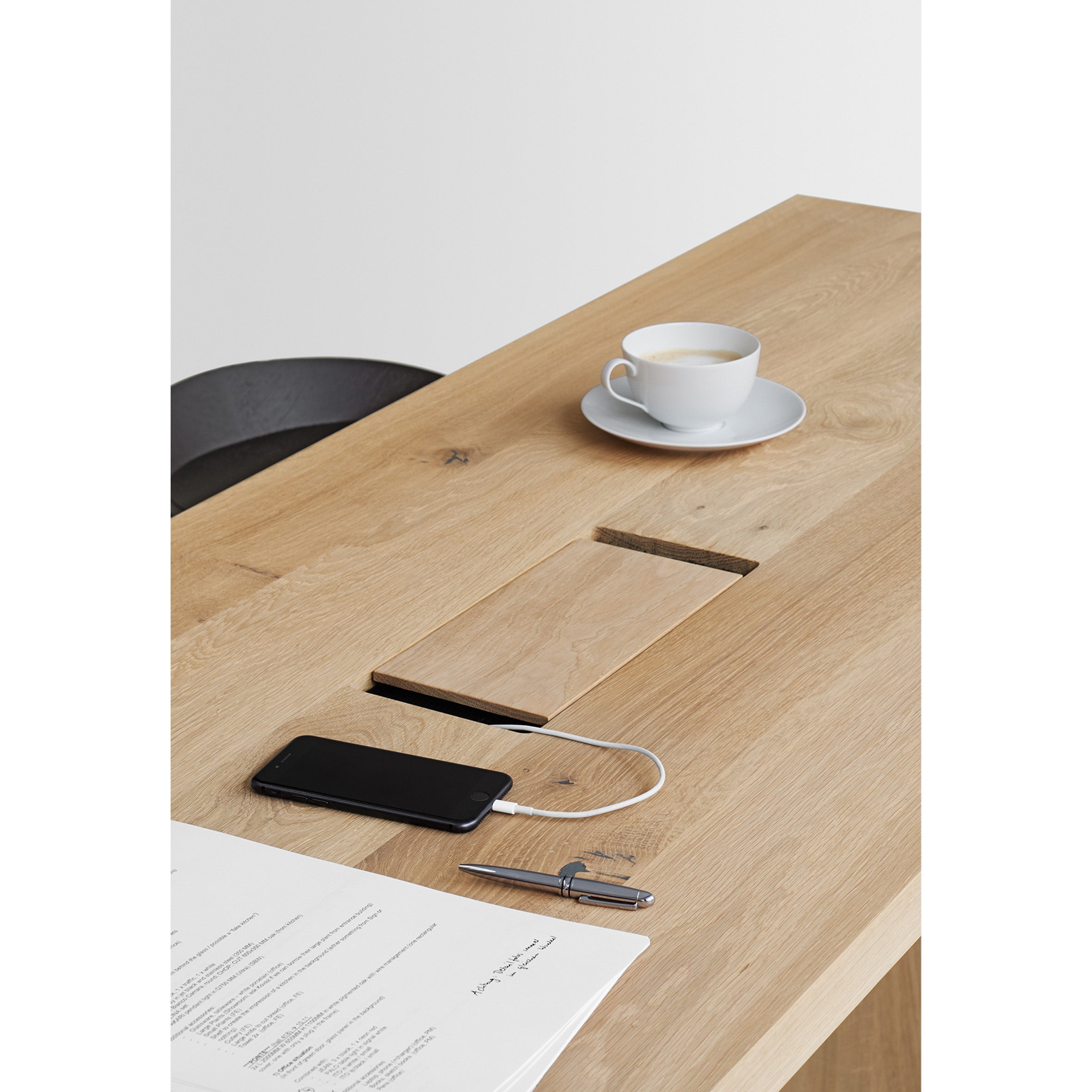The TA01 PONTE HIGH table by E15 features a white cup and saucer, a smartphone charging with a white cable, papers, a silver pen, and partially visible chair.