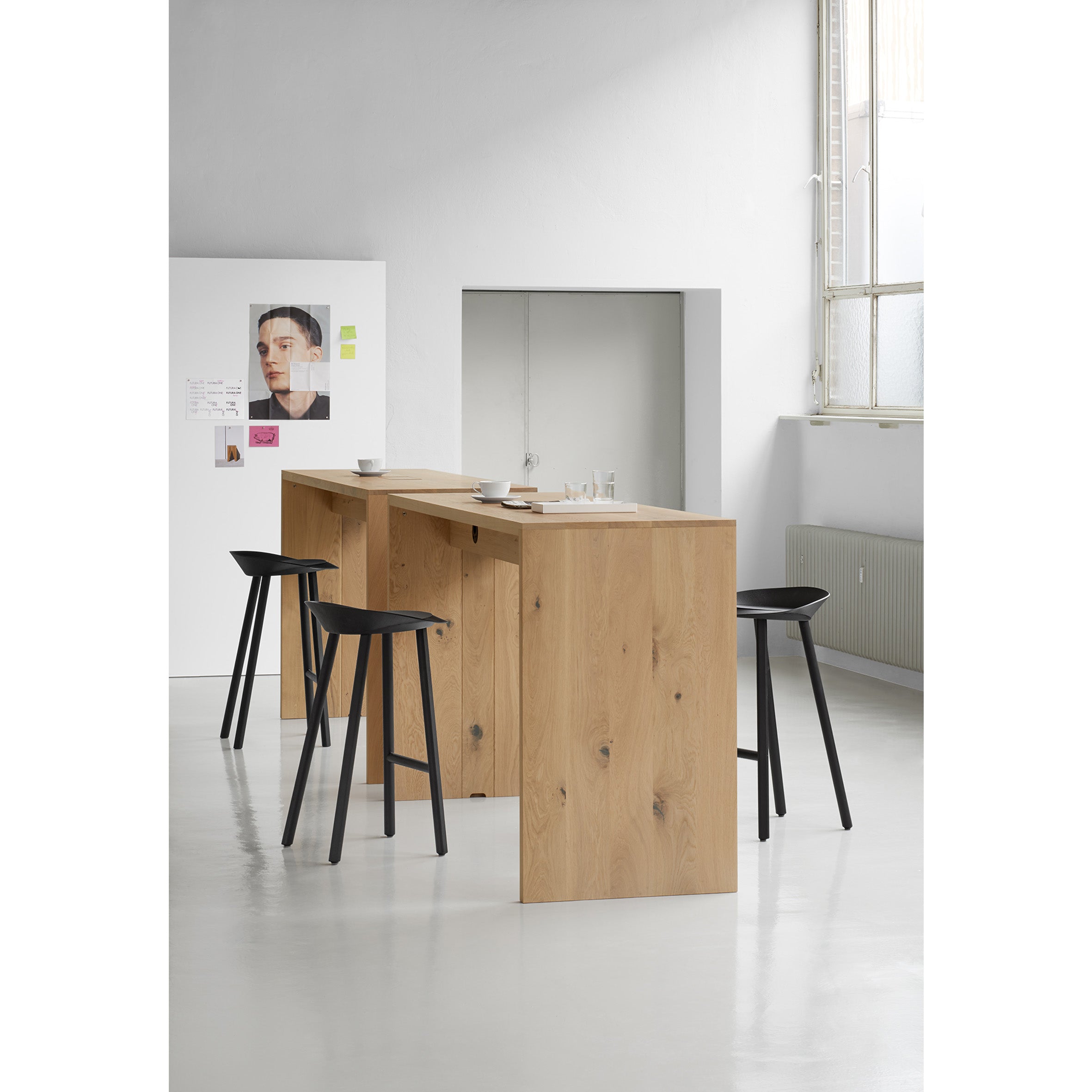 Bright and clean, this modern minimalist kitchen showcases a wooden island reminiscent of European walnut, paired with three black stools on a light floor. A wall with a window, radiator, and poster enhances the spaces elegance—a nod to E15s TA01 PONTE HIGH tables timeless design.