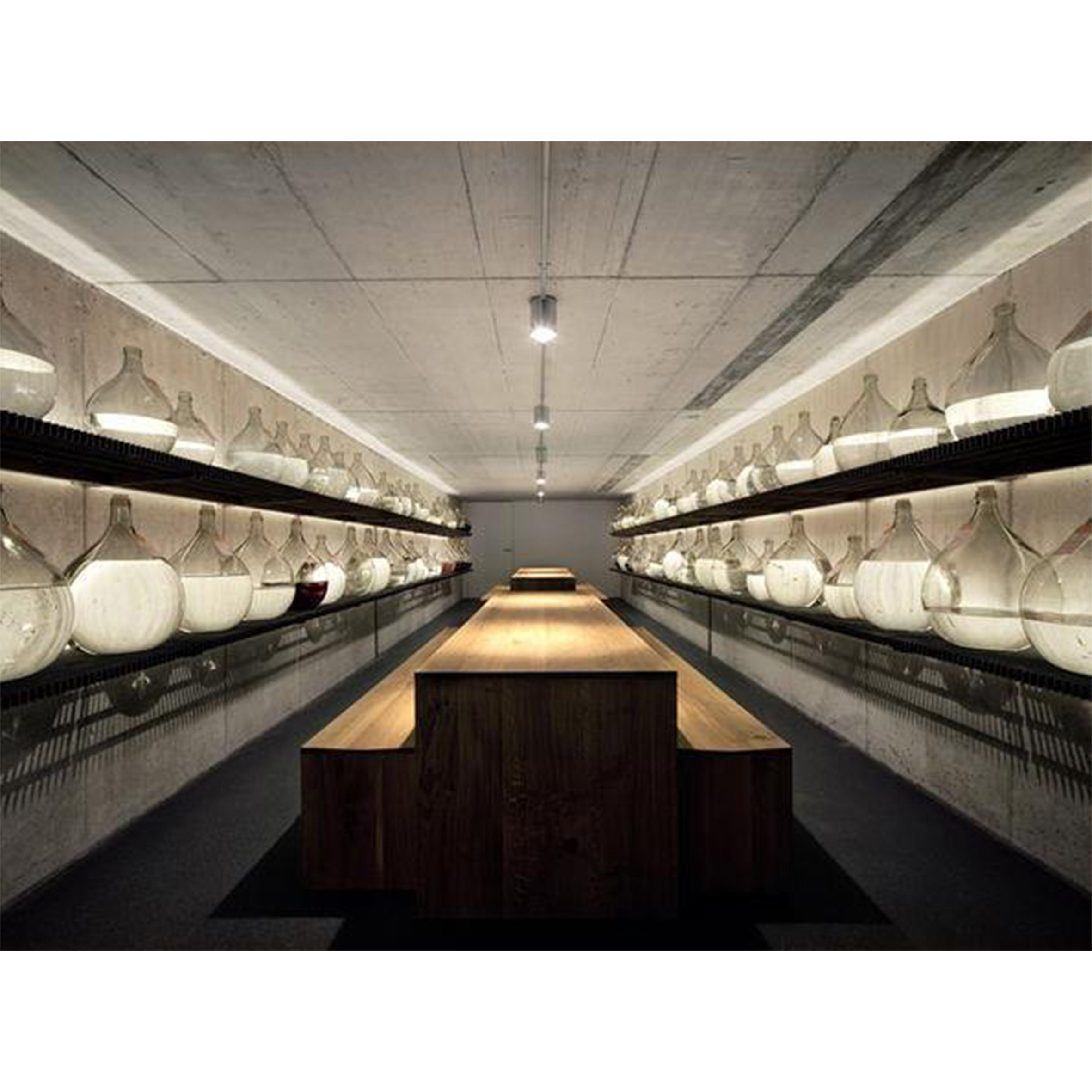 A modern, dimly lit cellar showcases glass jugs on shelves along both walls. A long TA01 PONTE conference table from E15 extends through the center, crafted from solid wood boards and embodying a sleek, symmetrical design with distinctive e15 style.