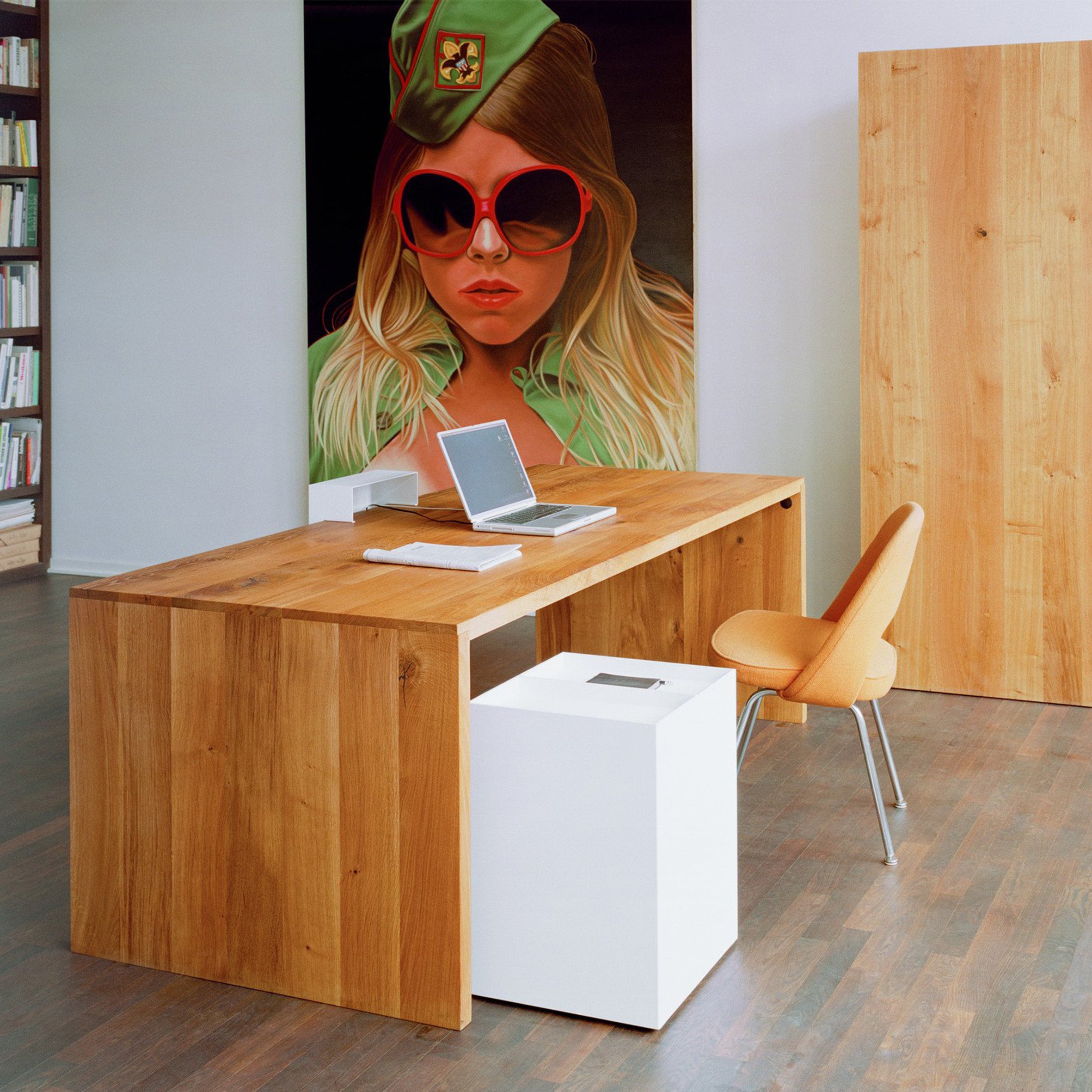 A modern office showcases an E15 TA01 PONTE wooden desk with a laptop and notepad, paired with an orange chair. A vibrant painting of a woman in sunglasses and a green cap accents the wall, while bookshelves line the left side.