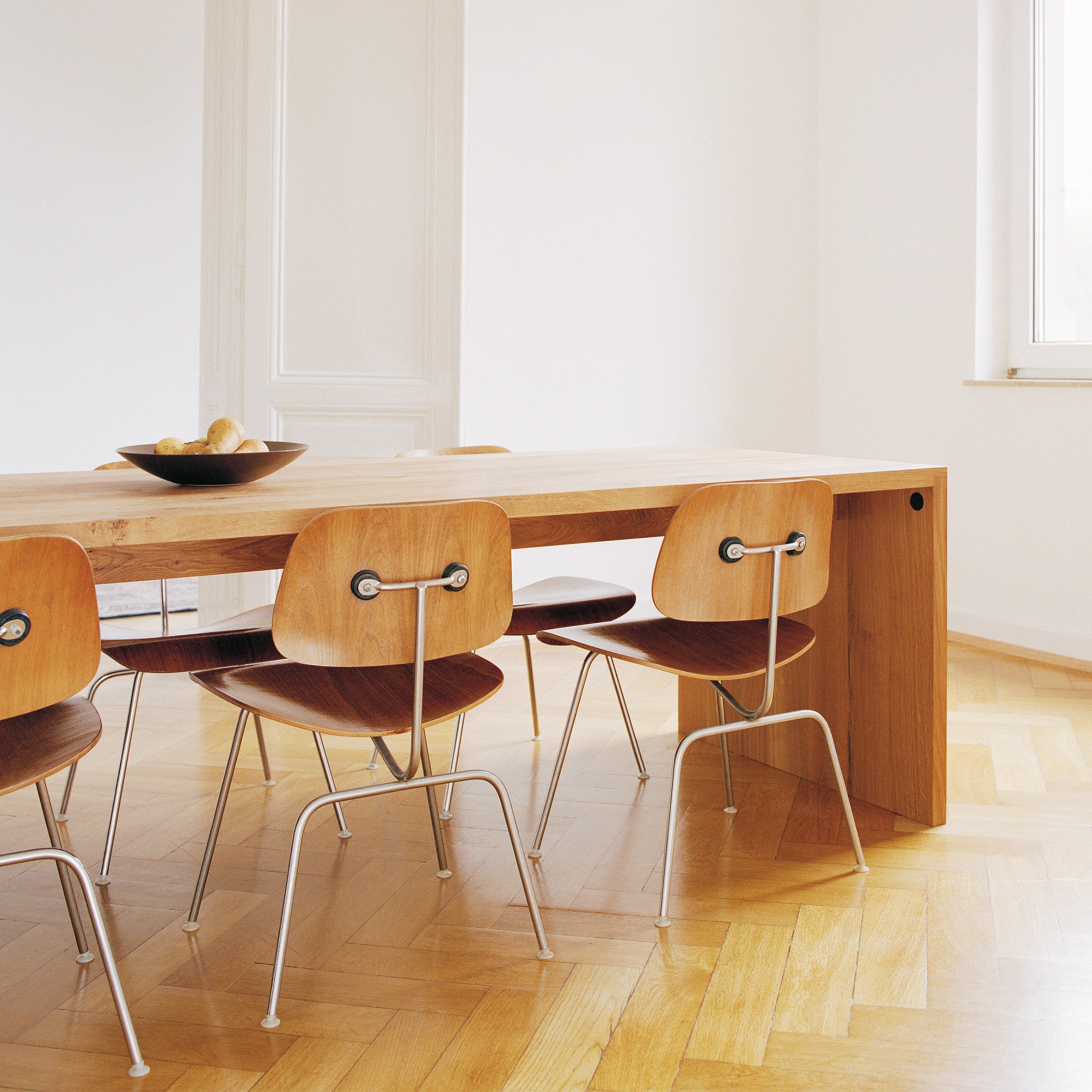 A modern dining room showcases an E15 TA01 PONTE wooden table with six chairs on a herringbone floor. A bowl of fruit adorns the table, and light streams through a right-side window, illuminating the space.