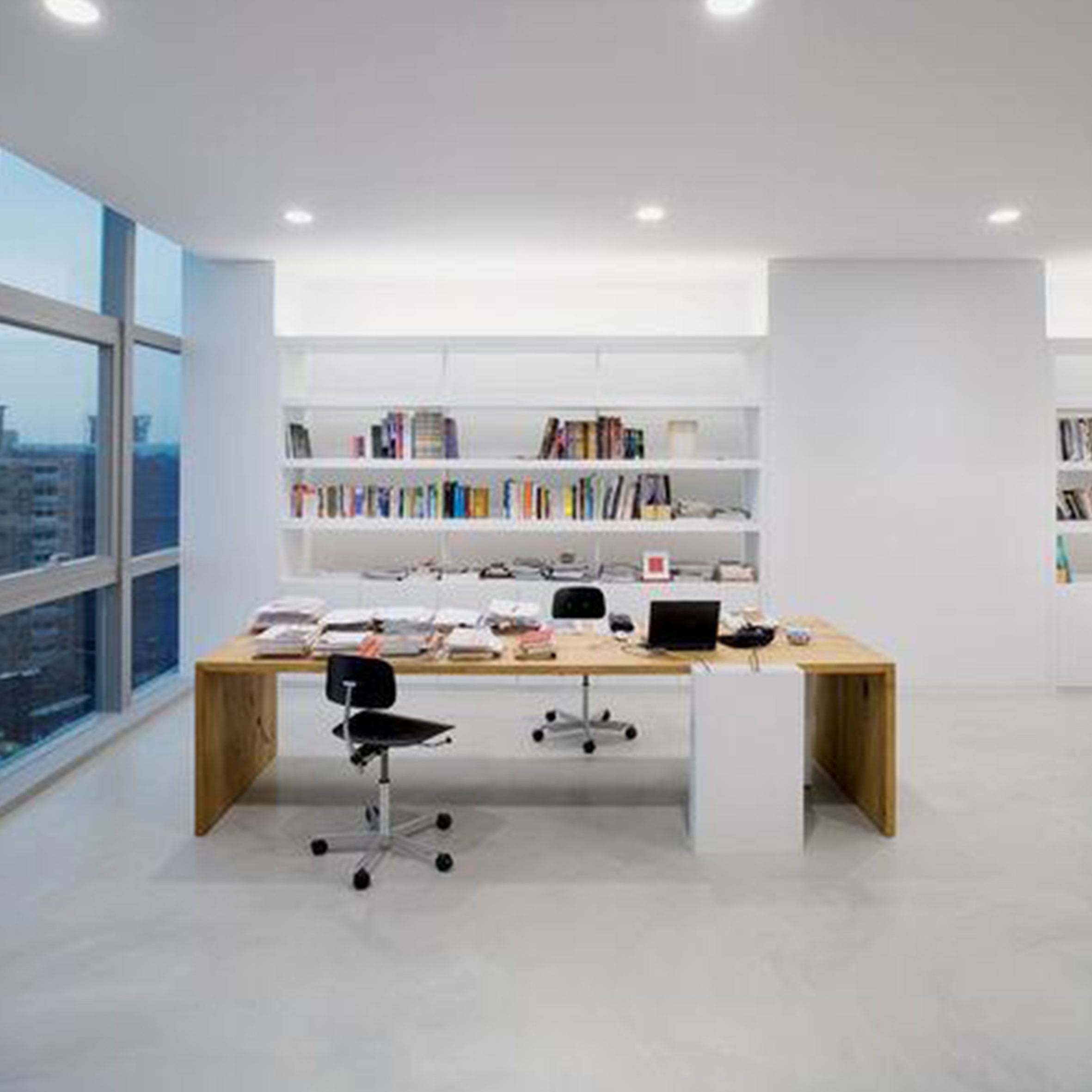 A modern office showcases the distinctive e15 TA01 PONTE design: a large wooden desk, similar to a conference table, faces a city-view window. The desk is filled with papers, a laptop, and stationery. Shelves packed with books line the far wall, brightly lit by recessed lighting.