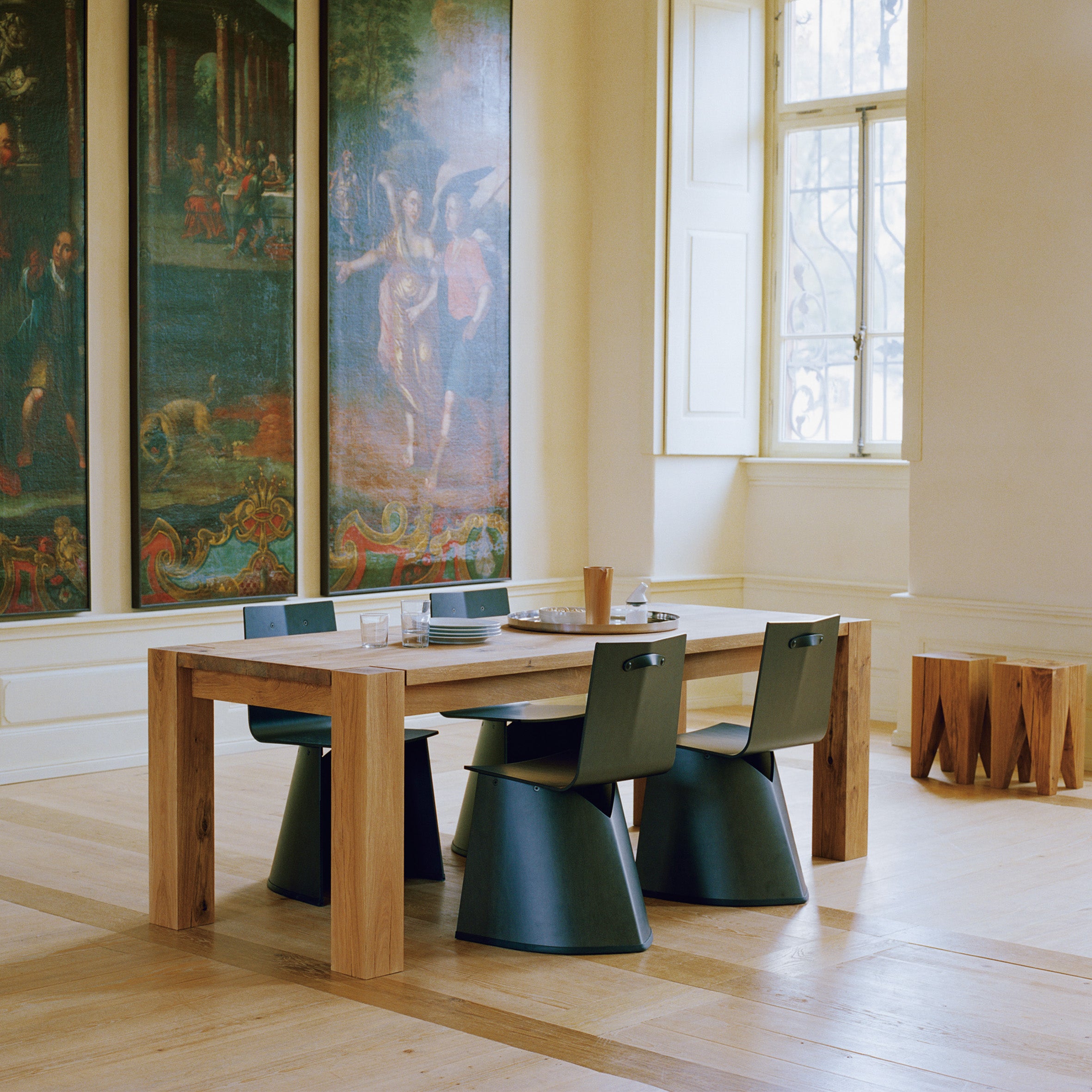 A modern dining area includes the E15 TA04 BIGFOOT, a classic solid wood table with four green cone-shaped chairs. Large art panels depicting classical scenes adorn the walls, and a tall window allows natural light to illuminate the wooden floor.