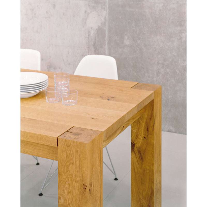 The TA04 BIGFOOT by E15, a classic-style wooden dining table with a natural finish, stands against a minimalist concrete wall. Its solid wood frame has sturdy legs and a smooth surface adorned with stacked white plates and clear glasses. Two white chairs are partially visible.