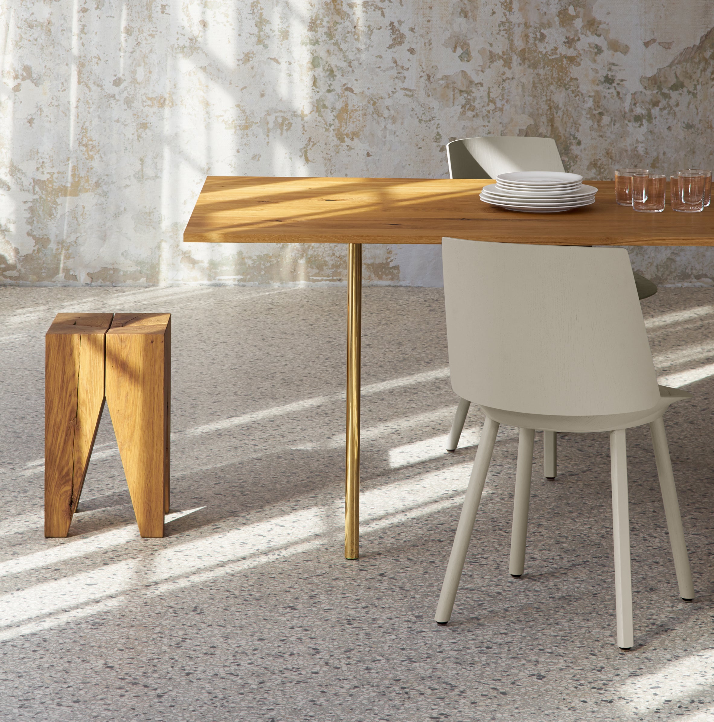The dining area features a TA14 ANTON walnut tabletop with a gold leg by E15, set with white plates and glasses. Two light-colored chairs and a wooden stool stand on speckled flooring against textured walls, creating a warm, modern ambiance.