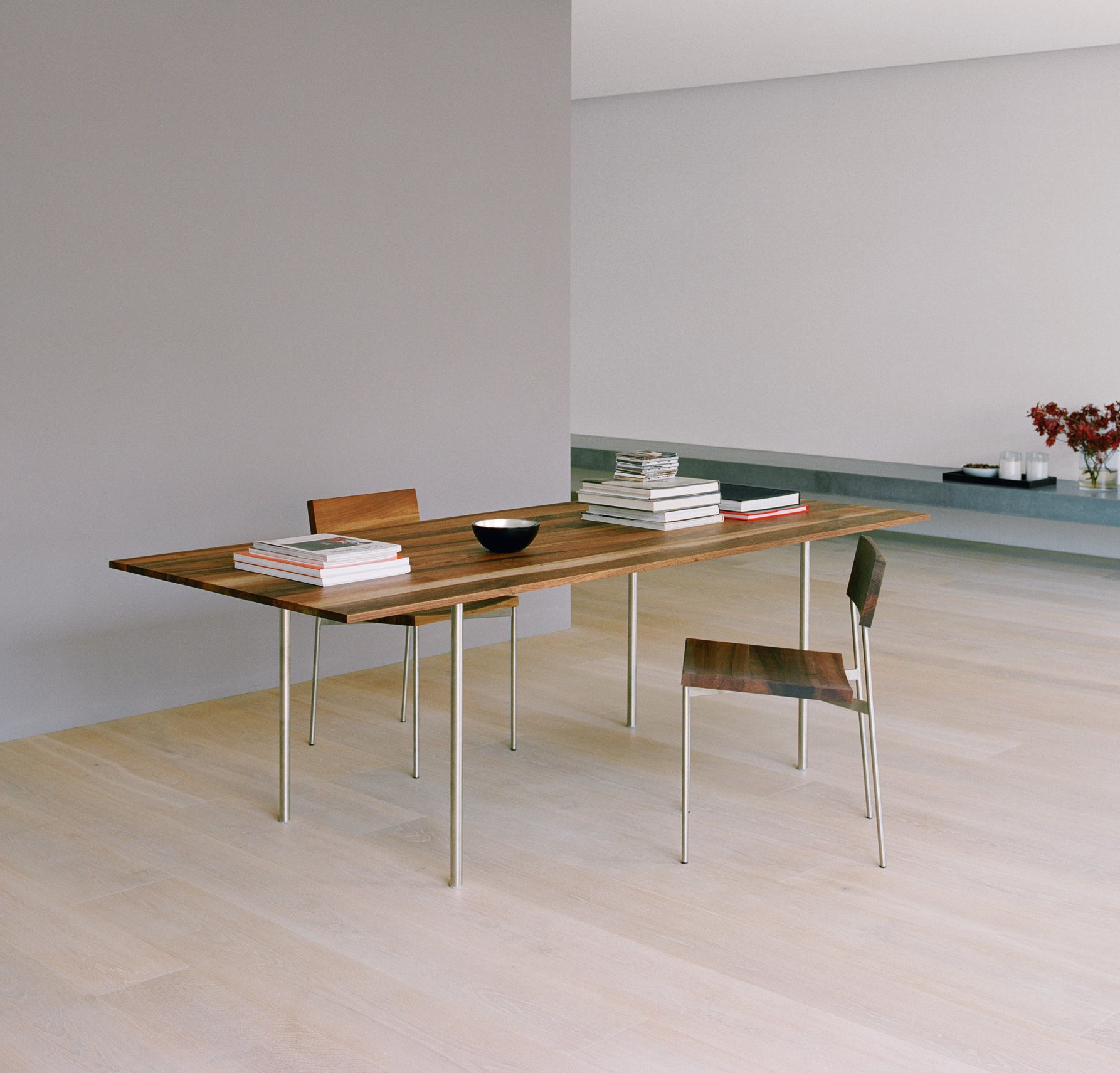 The minimalist interior showcases an E15 TA14 ANTON table with a solid wood top and sleek steel legs, paired with two matching chairs. The table features stacked books and a black bowl. The room includes light wooden flooring, gray walls, and a low shelf adorned with flowers in the background.