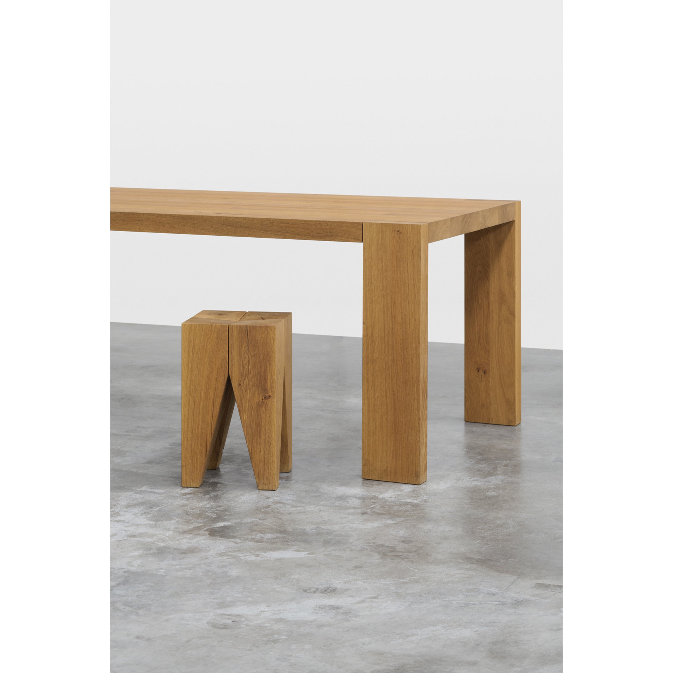 A minimalist wooden table crafted from solid wood with thick legs, similar to the classic TA17 LONDON design by E15, sits beside a small matching stool on a polished concrete floor. The plain white background highlights the furnitures natural finish and simple elegance.
