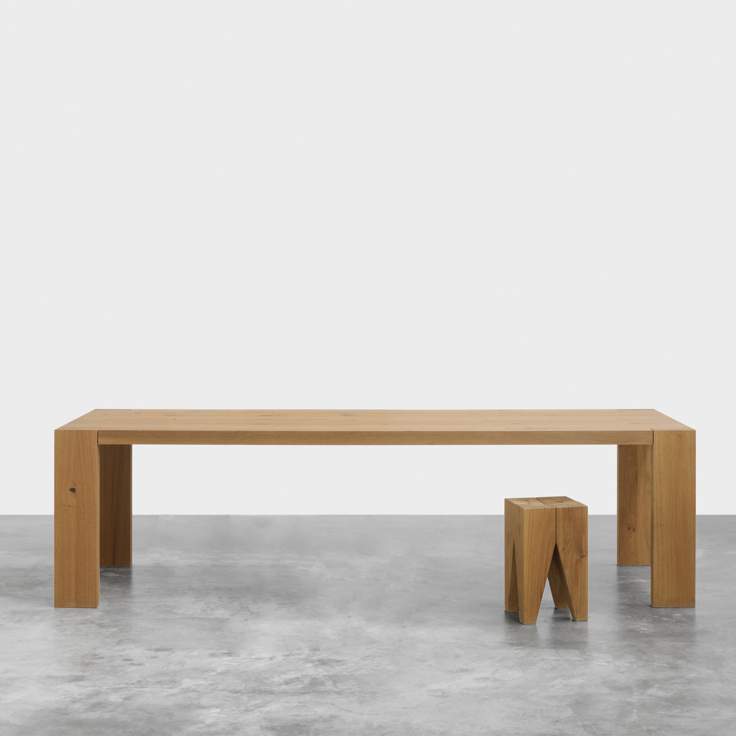 A minimalist interior with a long, rectangular solid wood table and a small wooden stool on polished concrete floors. The plain white walls enhance the clean design, capturing the classic E15 TA17 LONDON aesthetic.